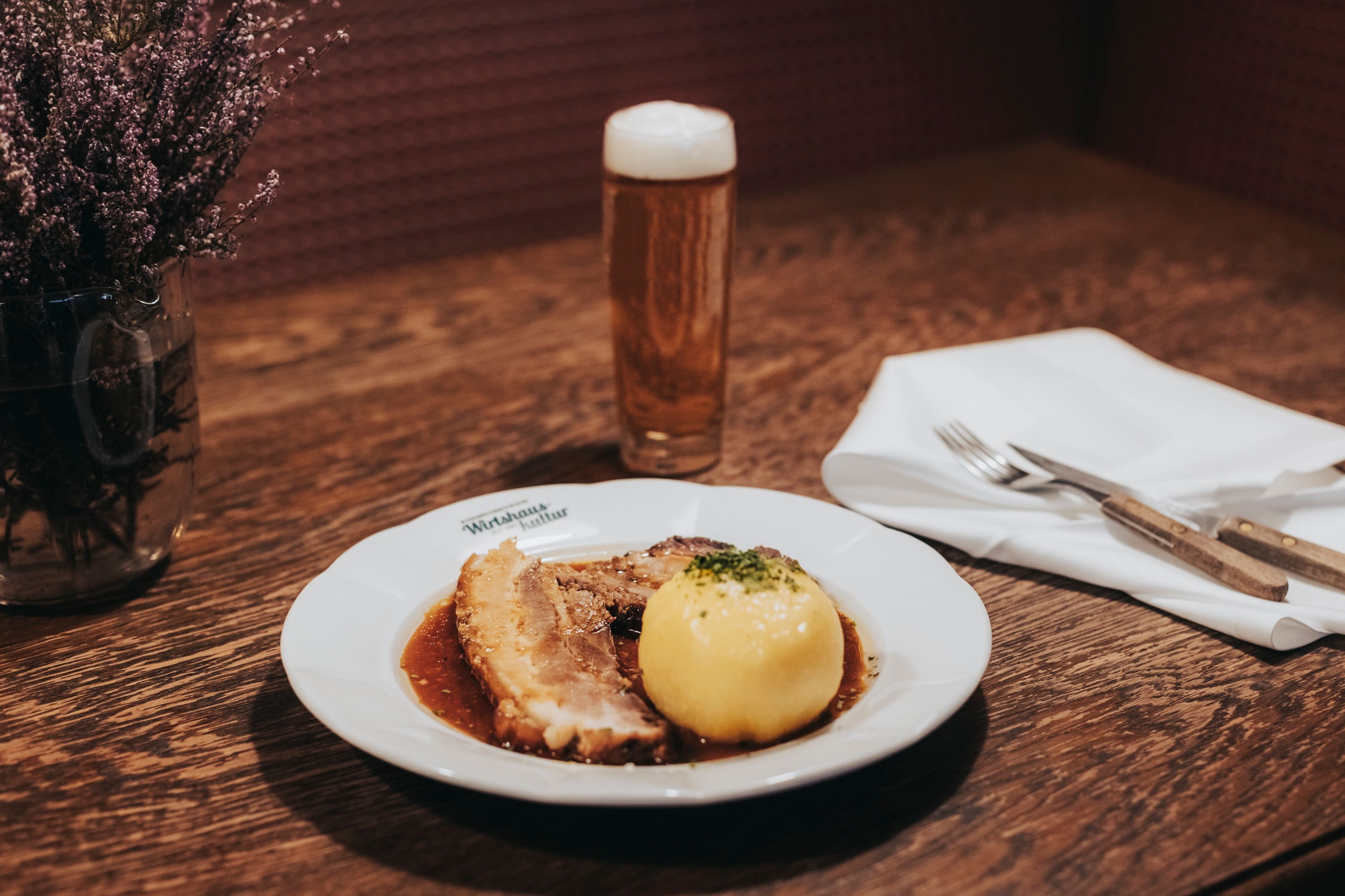 Ein Teller mit Schweinsbraten und Waldviertler Knödel, daneben ein Glas Bier auf einem Holztisch.