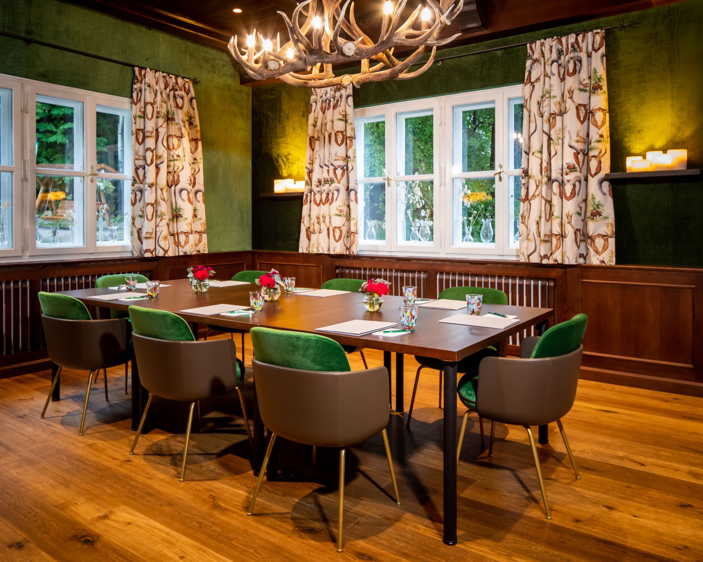 Ein Jagdzimmer mit Holztisch, grünen Stühlen und Geweihleuchter.