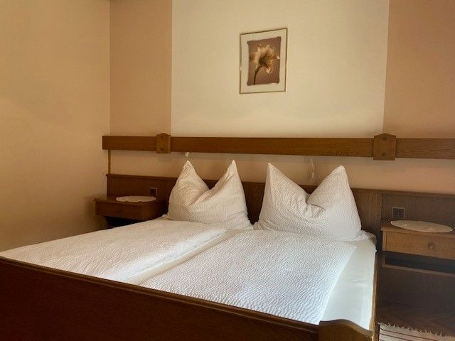 Ein gemütliches Doppelzimmer mit einem großen Bett, weißen Kissen und Decken, Holzmöbeln und einem Blumenbild an der Wand.