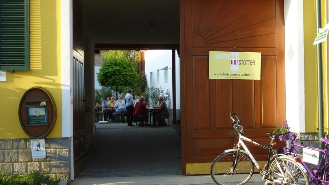 Eingang zu einem Weingut mit offenem Tor, durch das man Menschen an Tischen sitzen sieht. Ein Fahrrad steht davor.