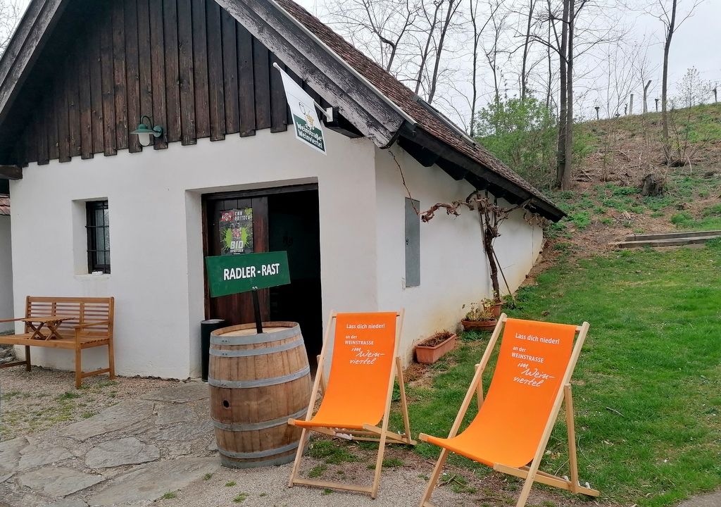 Kleines weißes Gebäude mit Holzdach, zwei orange Liegestühle und ein Holzfass davor, umgeben von Bäumen und Gras.