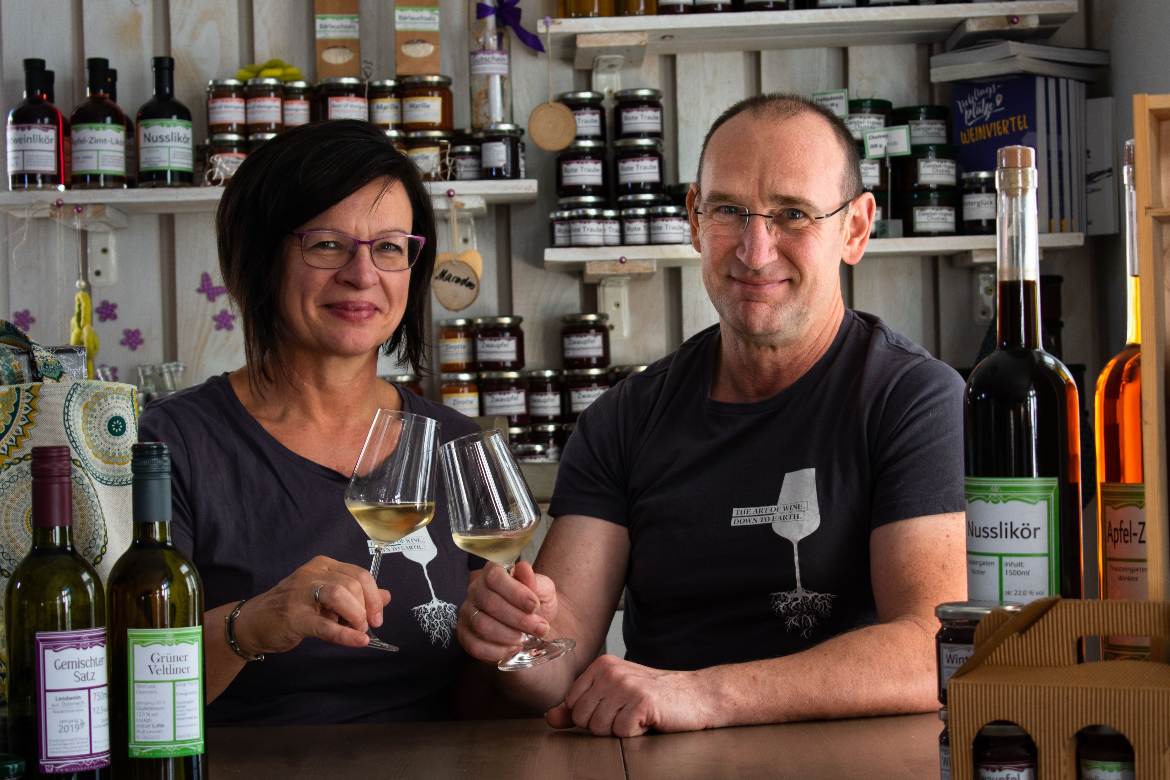Ein Mann und eine Frau sitzen an einem Tisch in einem Hofladen, halten Weingläser und lächeln. Im Hintergrund sind Regale mit Produkten zu sehen.
