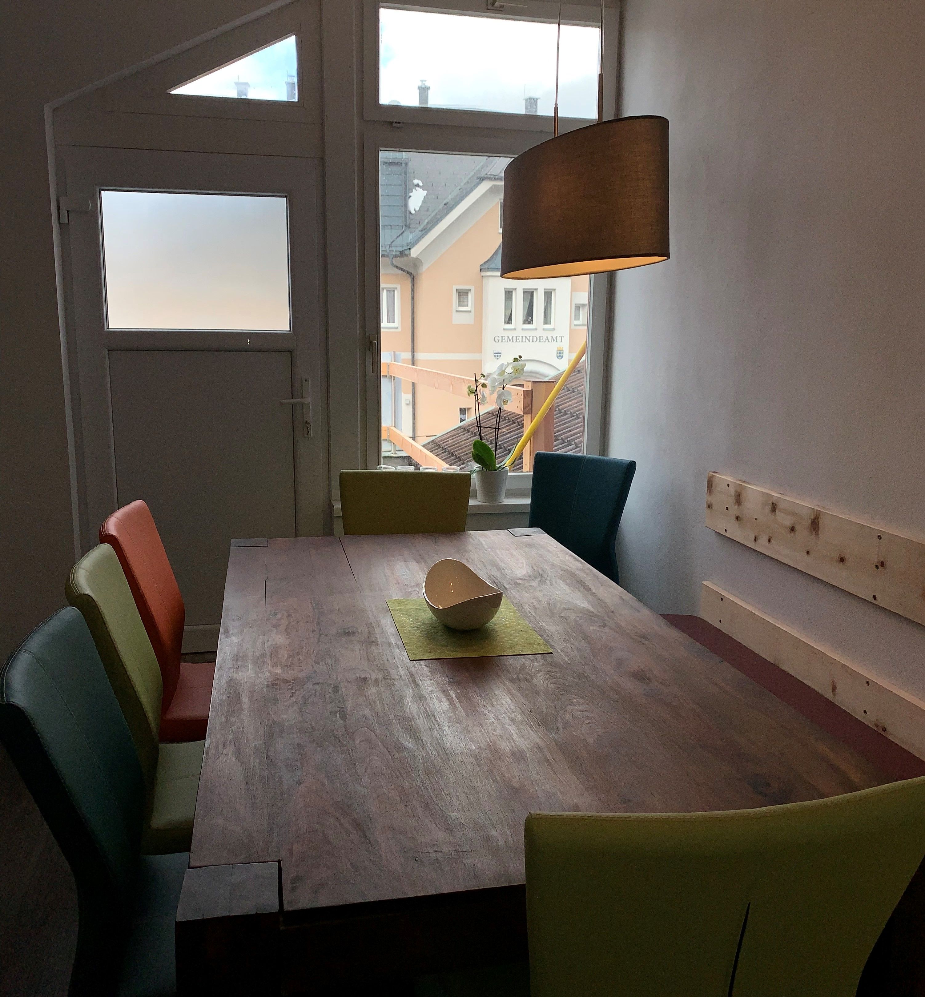 Essbereich mit Holztisch, bunten Stühlen und Hängelampe, Fenster mit Blick auf Gebäude.
