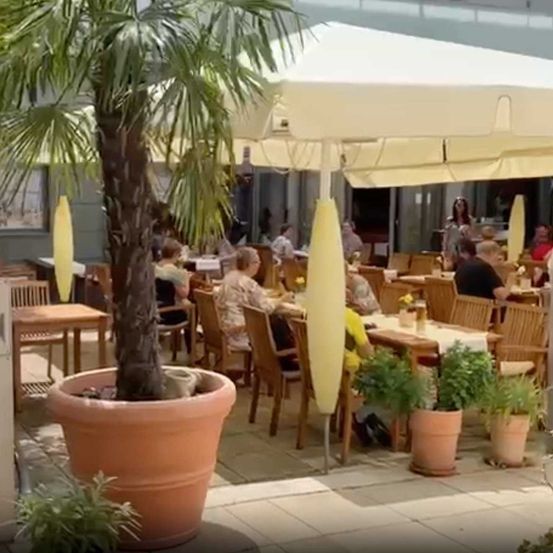 Ein Außenbereich eines Cafés mit Holztischen und -stühlen, umgeben von Pflanzen und einem großen Sonnenschirm.