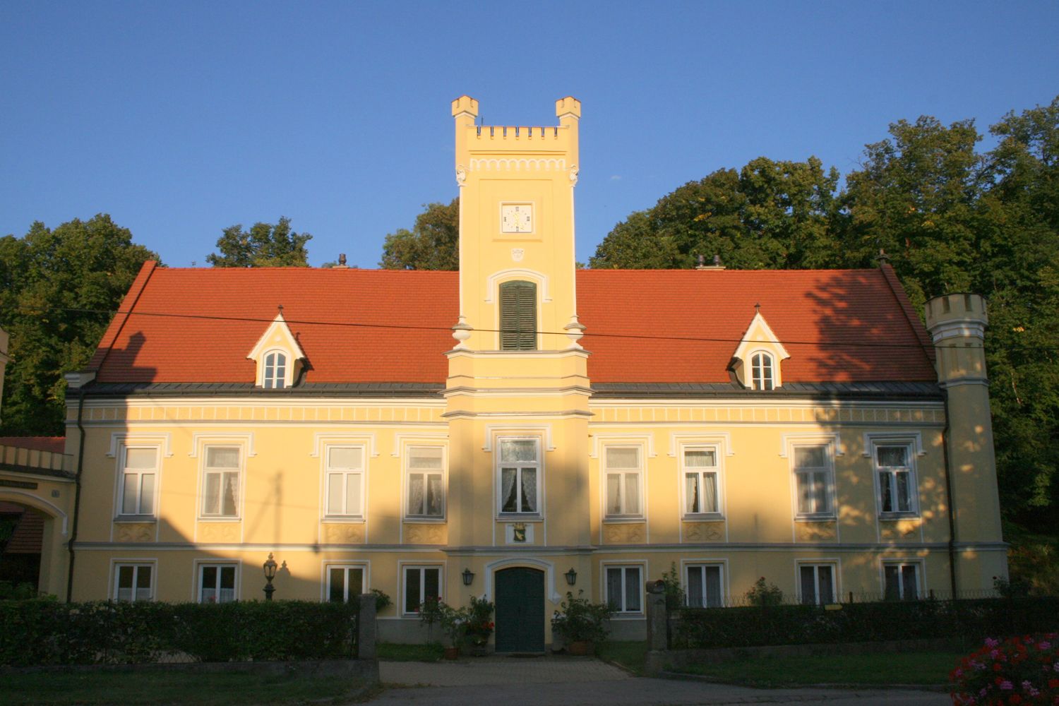 Gelbes Schloss mit rotem Dach und Turm, umgeben von Bäumen.