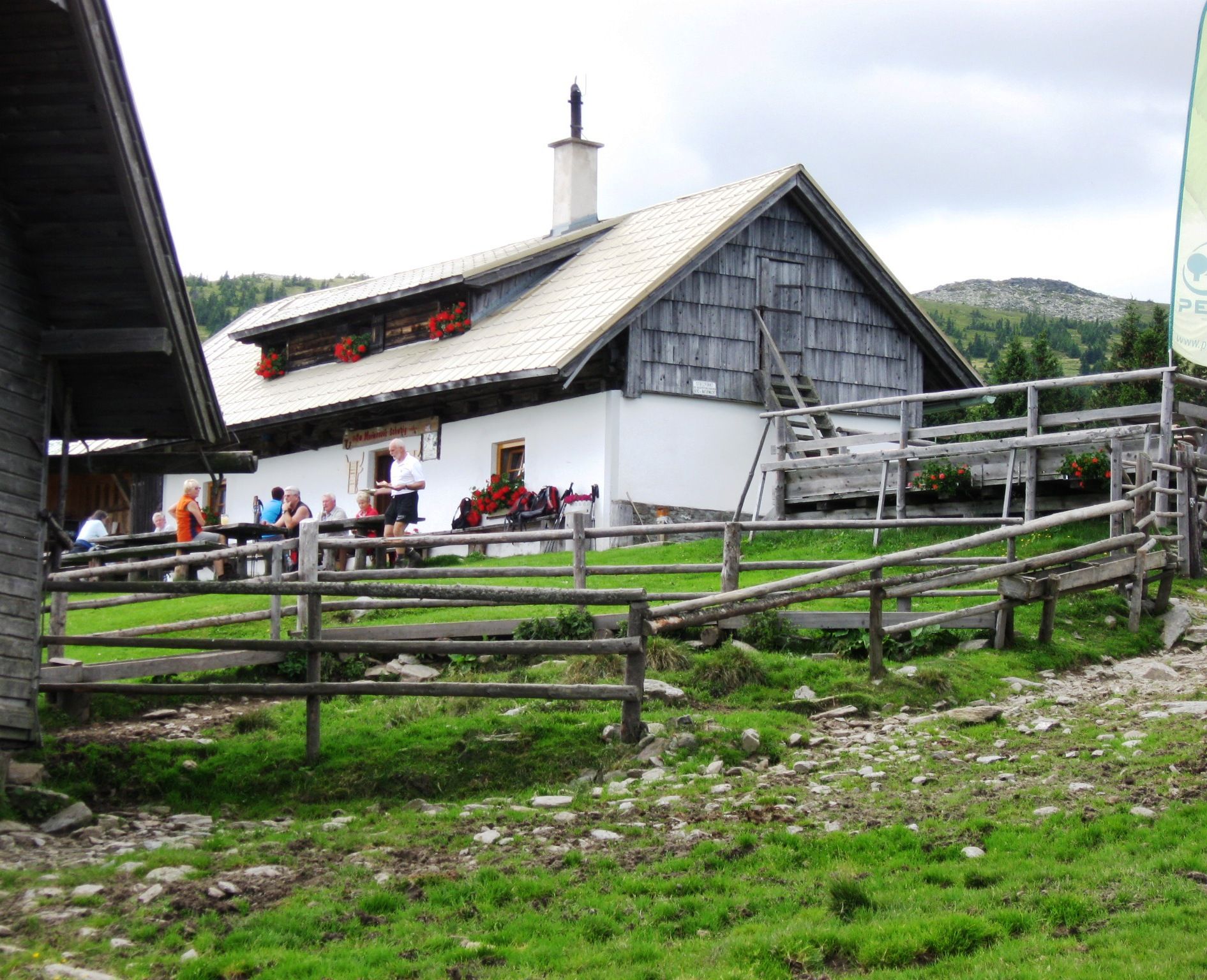 Almhütte mit Menschen auf einer Wiese, umgeben von Holzzäunen.