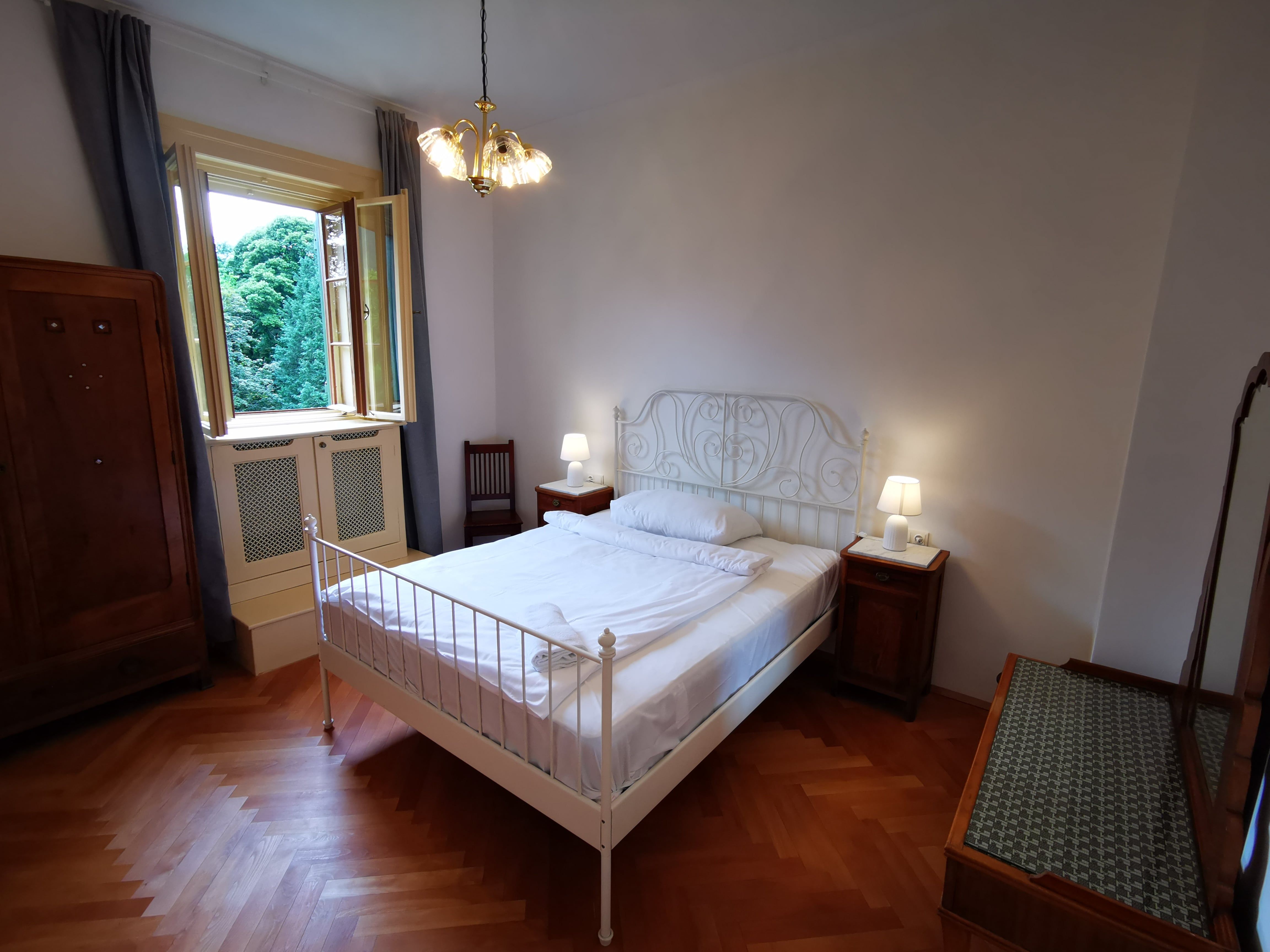 Schlafzimmer mit Doppelbett, Nachttischen und offenem Fenster mit Blick auf Bäume.