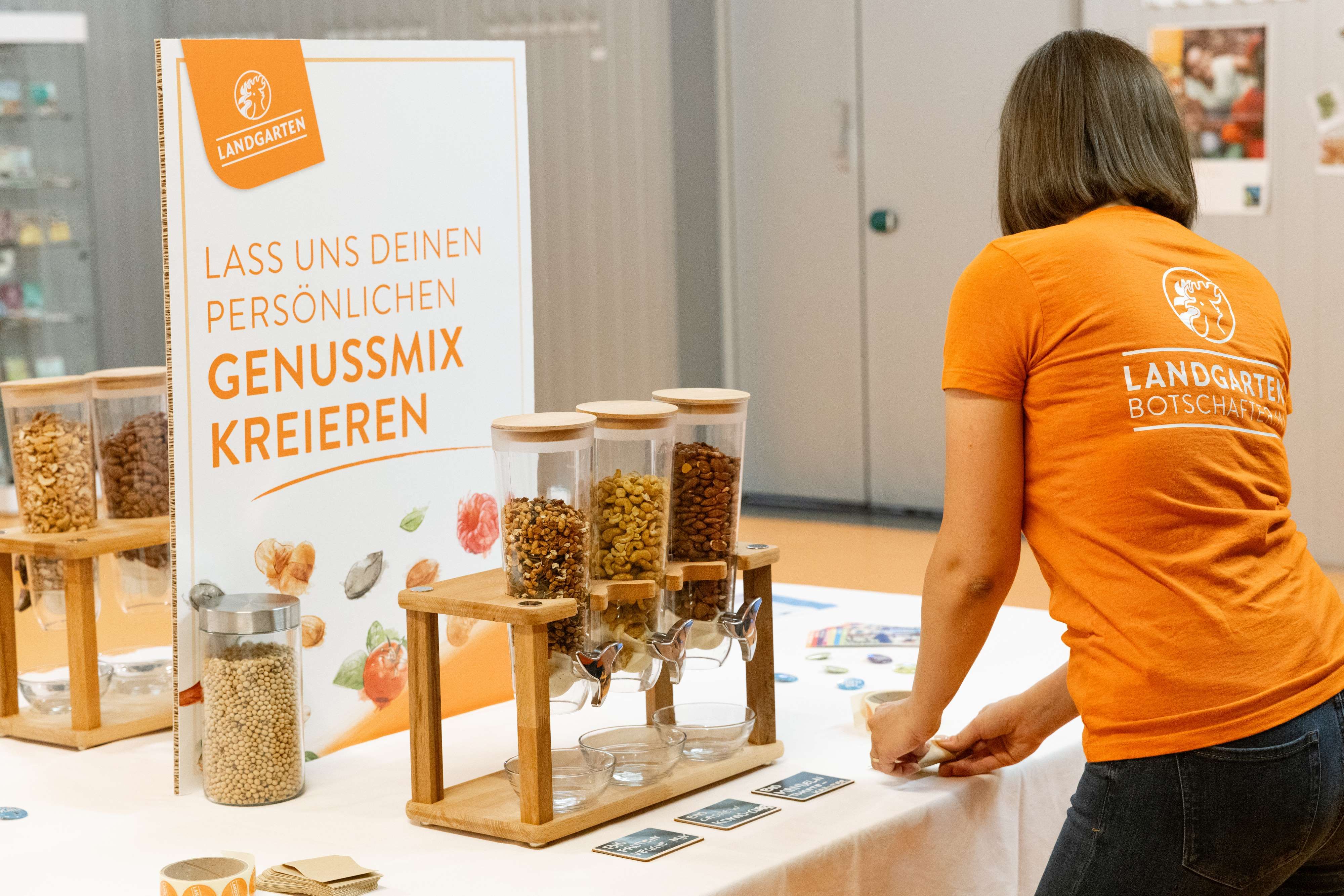 Eine Person in einem orangefarbenen T-Shirt steht an einem Tisch mit Nuss- und Snackspendern. Ein Schild wirbt für einen persönlichen Genussmix.