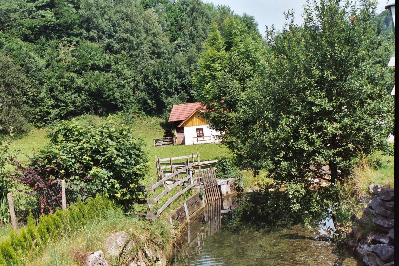 Eine kleine Hütte mit rotem Dach steht in einer grünen Landschaft neben einem Bach und Bäumen.