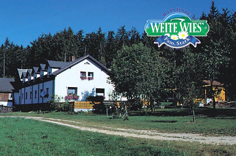 Ein weißes Bauernhaus mit Blumen vor einem Wald, darüber das Logo 'Biohof Weite Wies Fam. Stark'.