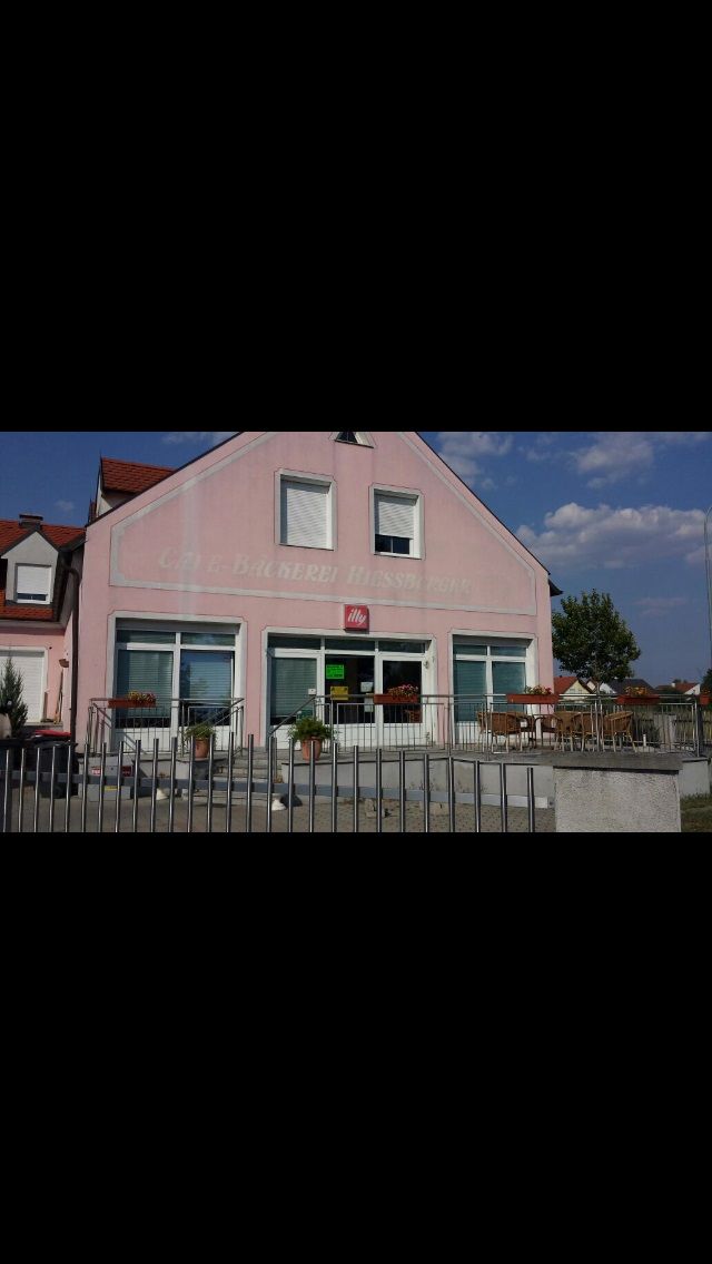 Außenansicht eines rosa Gebäudes mit der Aufschrift 'Bäckerei-Café Hiessberger'. Vor dem Gebäude stehen Tische und Stühle.