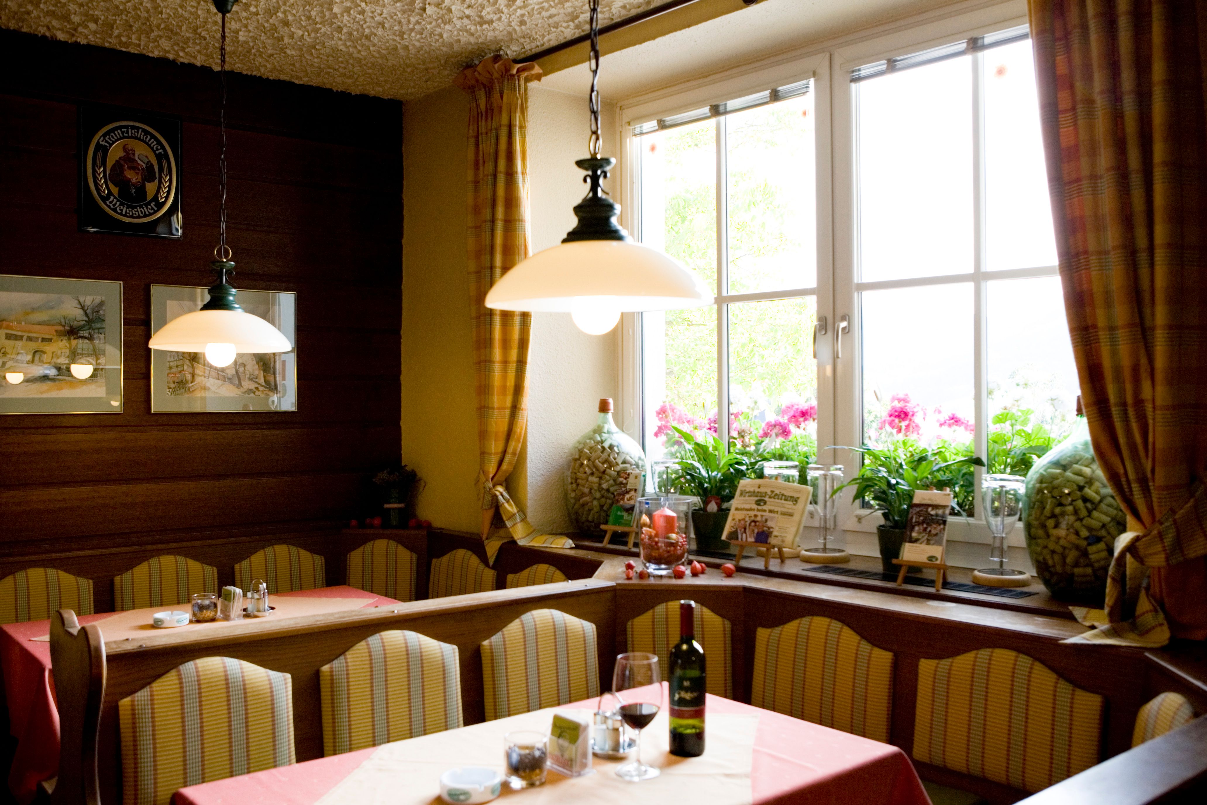 Eine gemütliche Gaststube mit Bänken, Tischen, Lampen und einem großen Fenster mit Vorhängen.