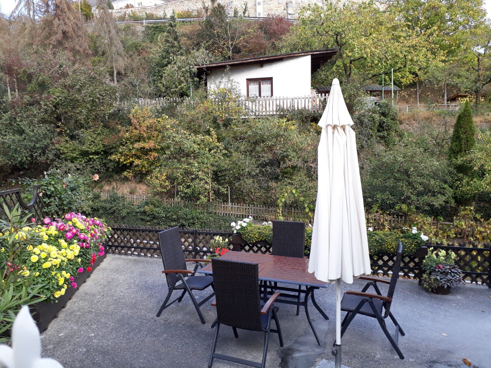 Terrasse mit Tisch, Stühlen und Sonnenschirm, umgeben von Blumen und Bäumen.