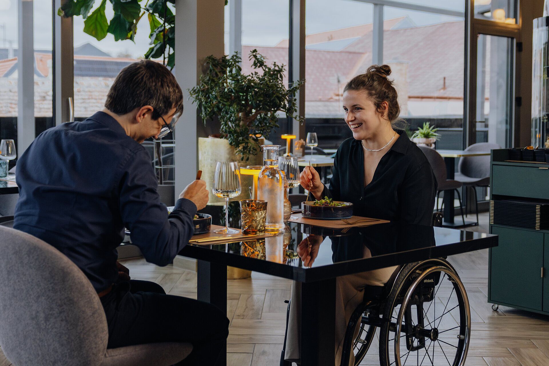 Eine Frau im Rollstuhl und ein Mann sitzen in einem Restaurant an einem Tisch gegenüber. Sie genießen ein kulinarisches Gericht und unterhalten sich.