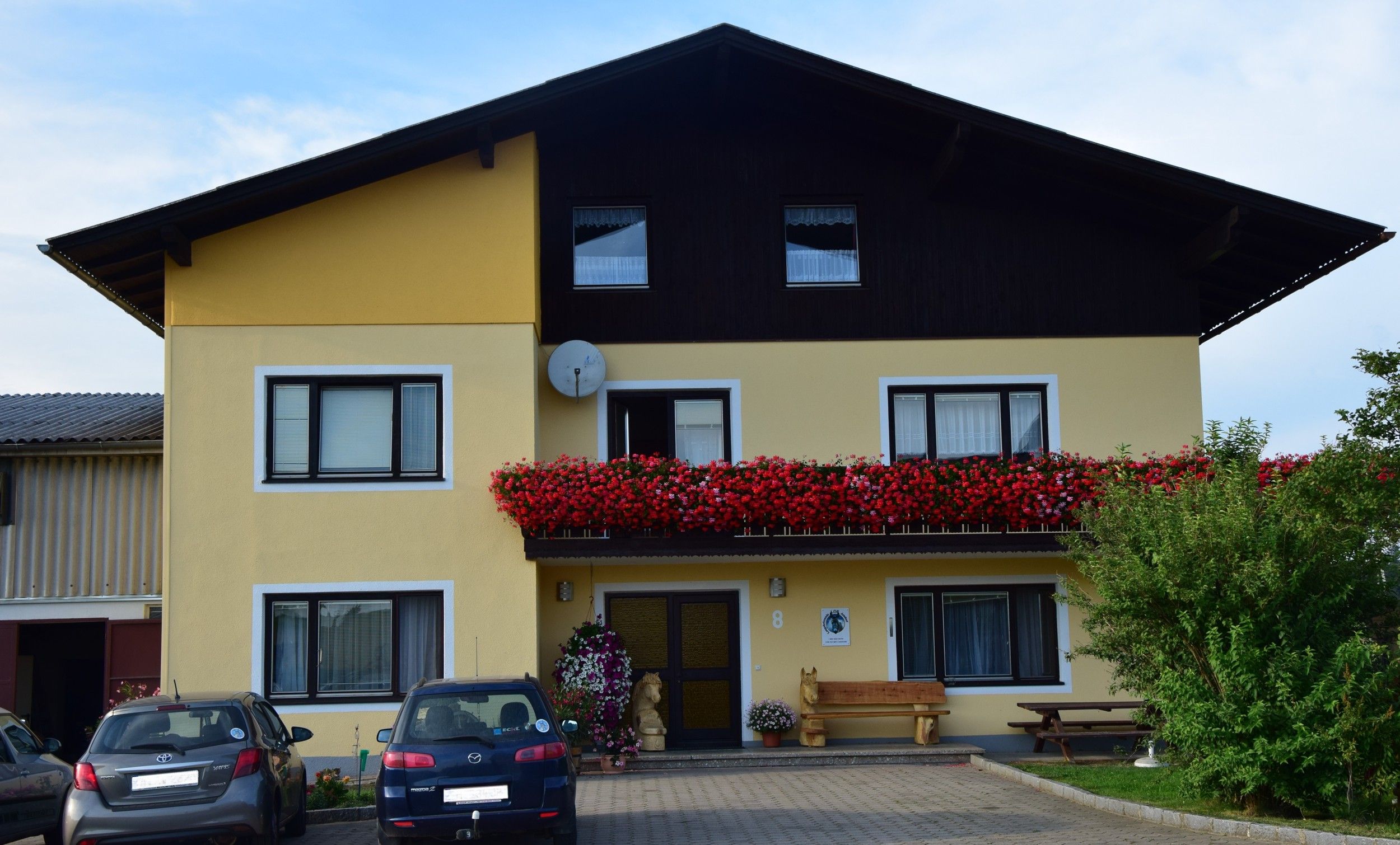 Außenansicht eines gelben Hauses mit Balkon voller roter Blumen und geparkten Autos davor.