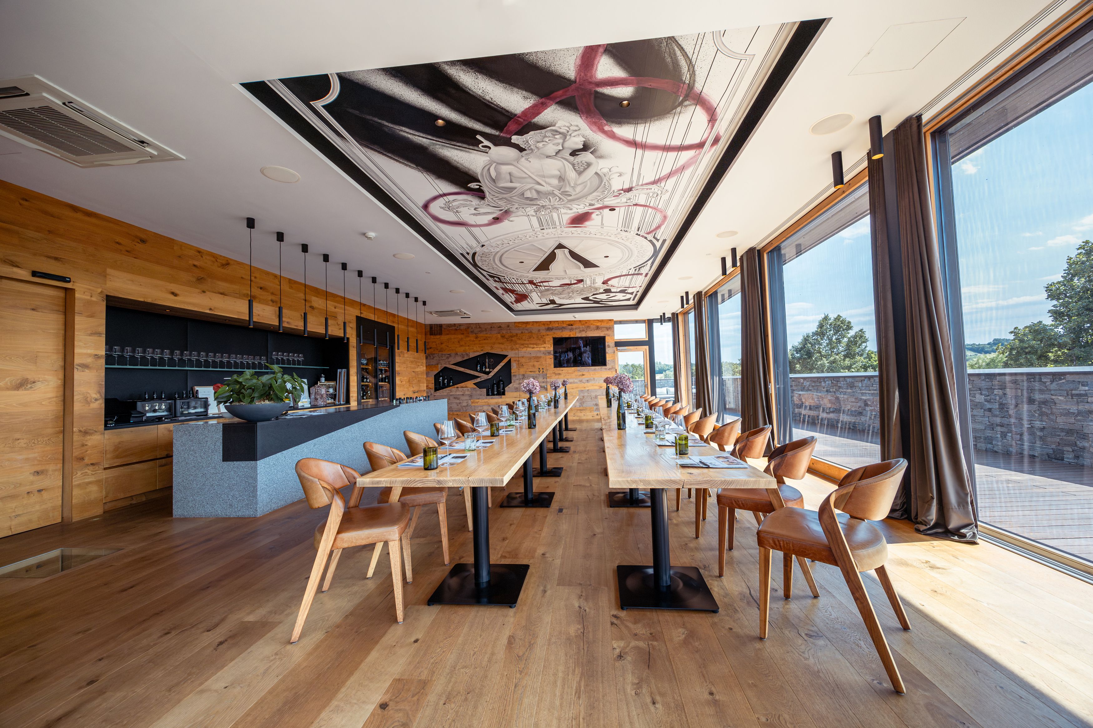 Innenansicht eines modernen Restaurants mit langen Holztischen, Ledersesseln und großen Fenstern.