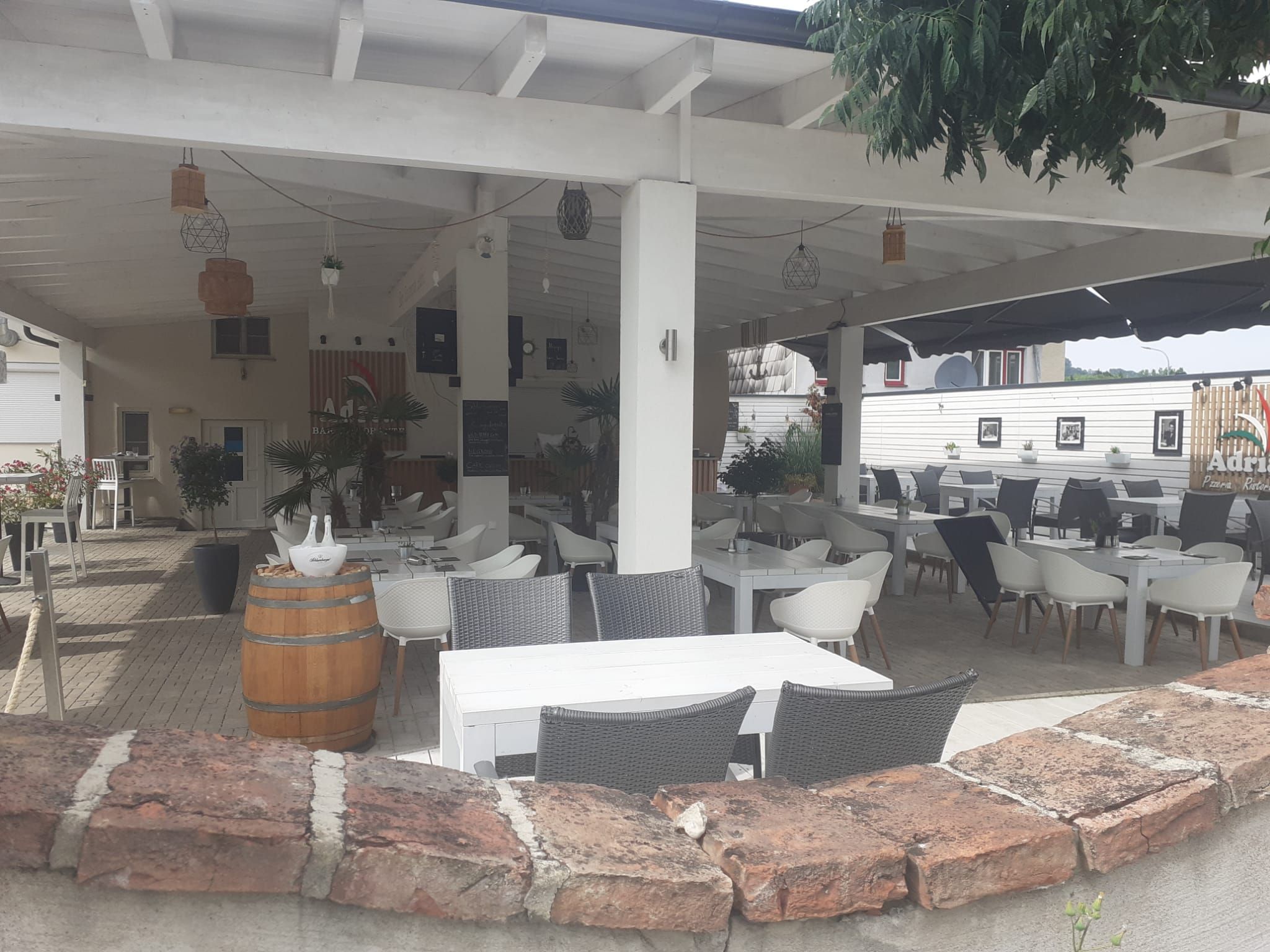 Ein überdachter Gastgarten mit weißen Tischen und Stühlen, umgeben von Pflanzen und Dekorationen.