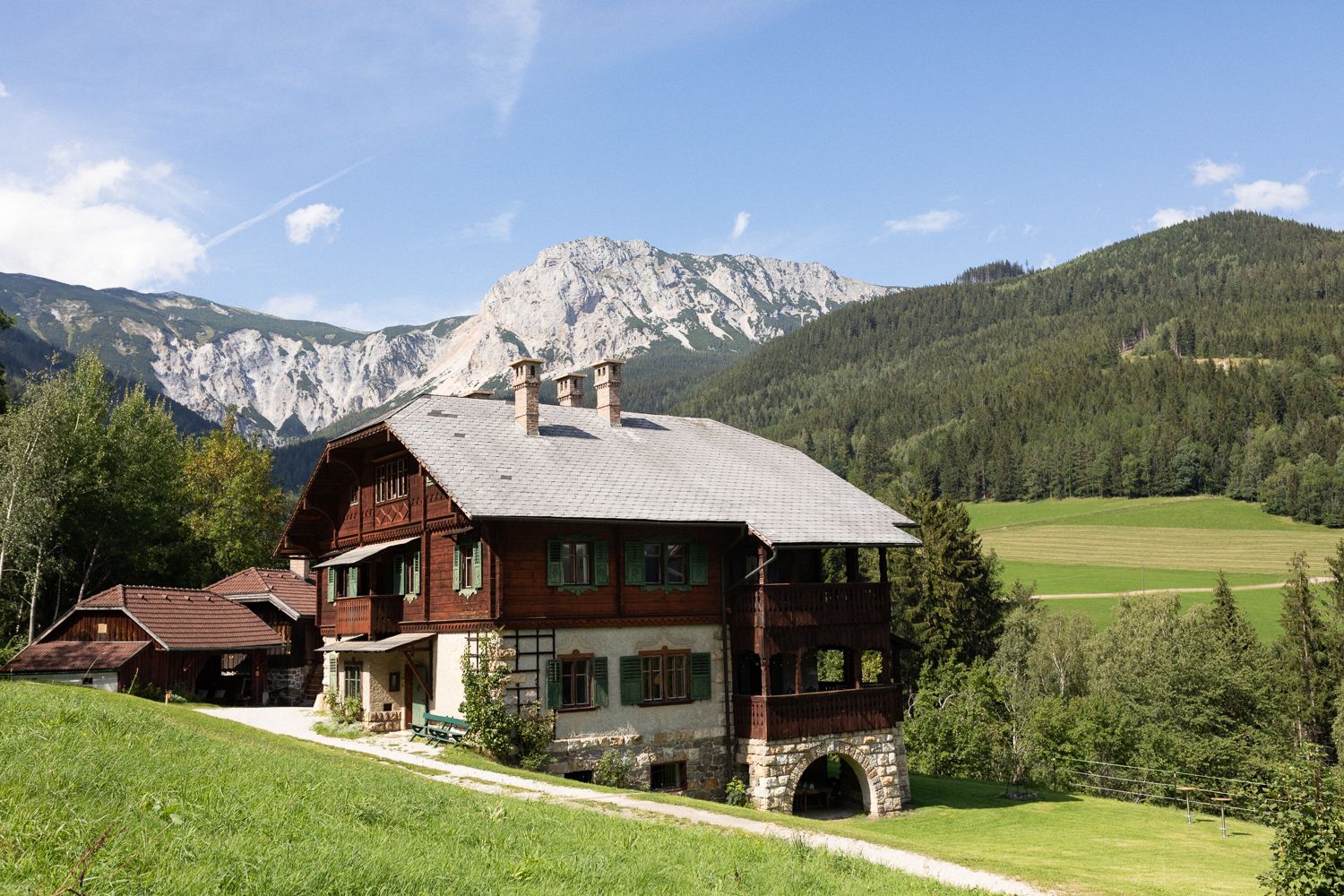 Ein traditionelles Holzhaus in einer bergigen Landschaft mit Wiesen und Wäldern im Hintergrund.