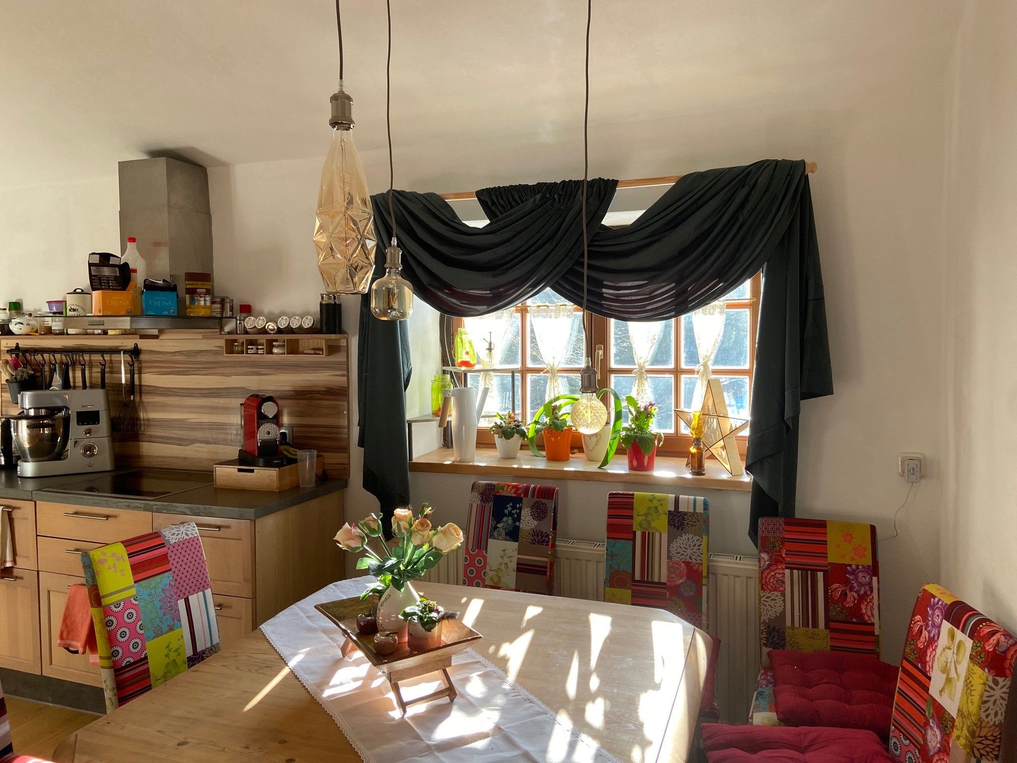 Essbereich mit Holztisch, bunten Stühlen, Pflanzen auf der Fensterbank und Hängelampen.