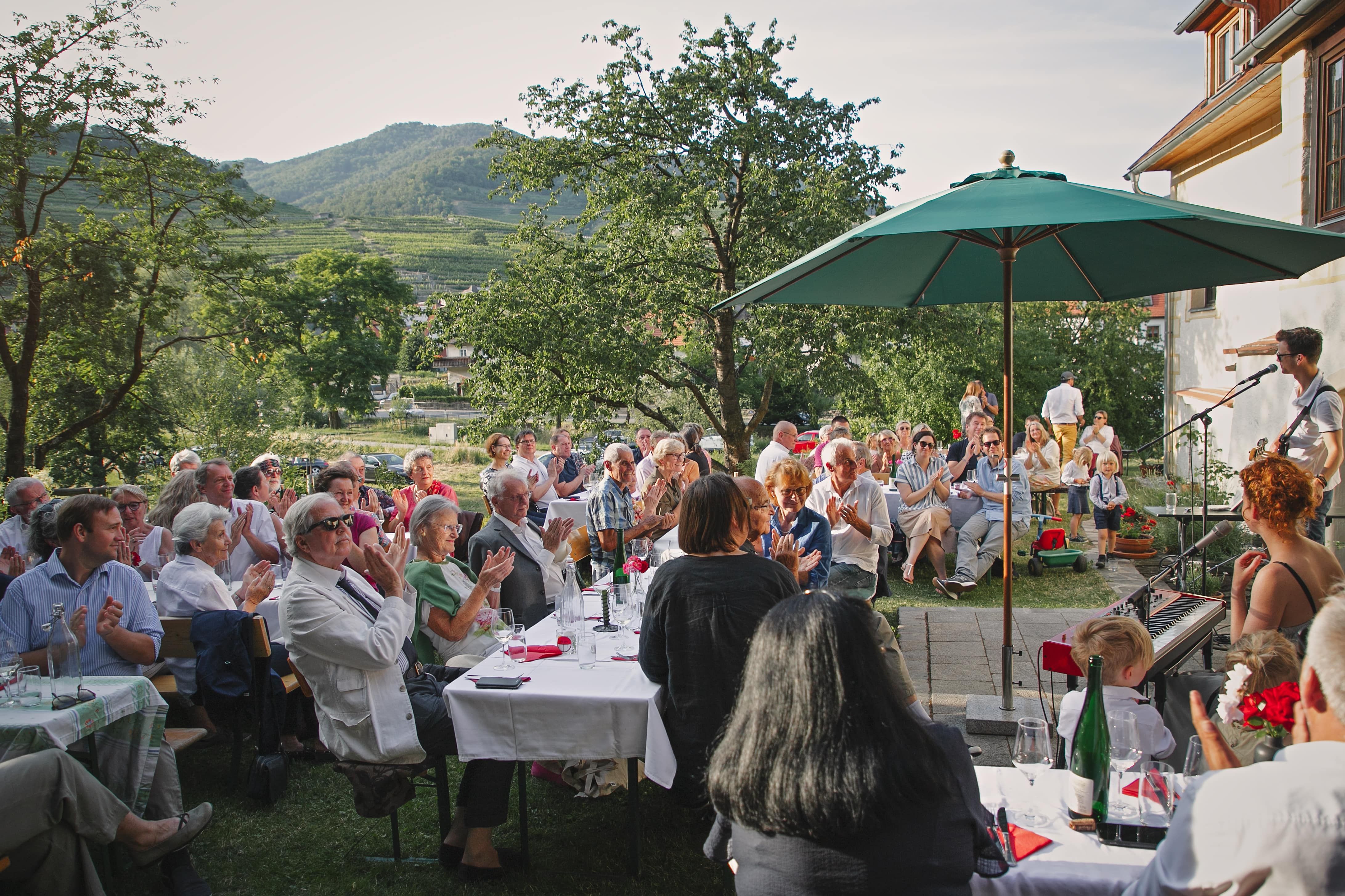 Menschen sitzen im Freien bei einer Veranstaltung mit Musik, umgeben von Bäumen und Bergen.