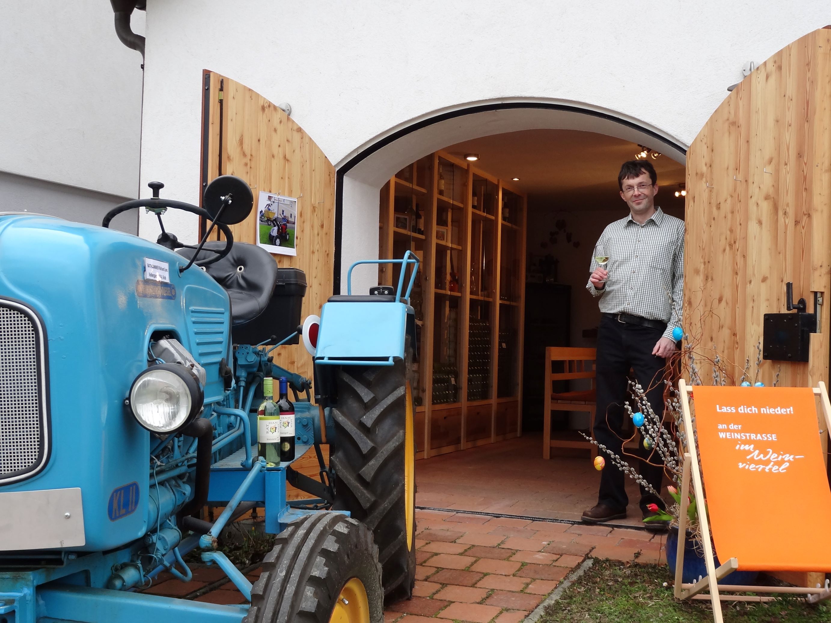 Ein Mann steht mit einem Weinglas in der Hand vor einem Gebäude mit offenen Holztüren. Ein blauer Traktor steht daneben.