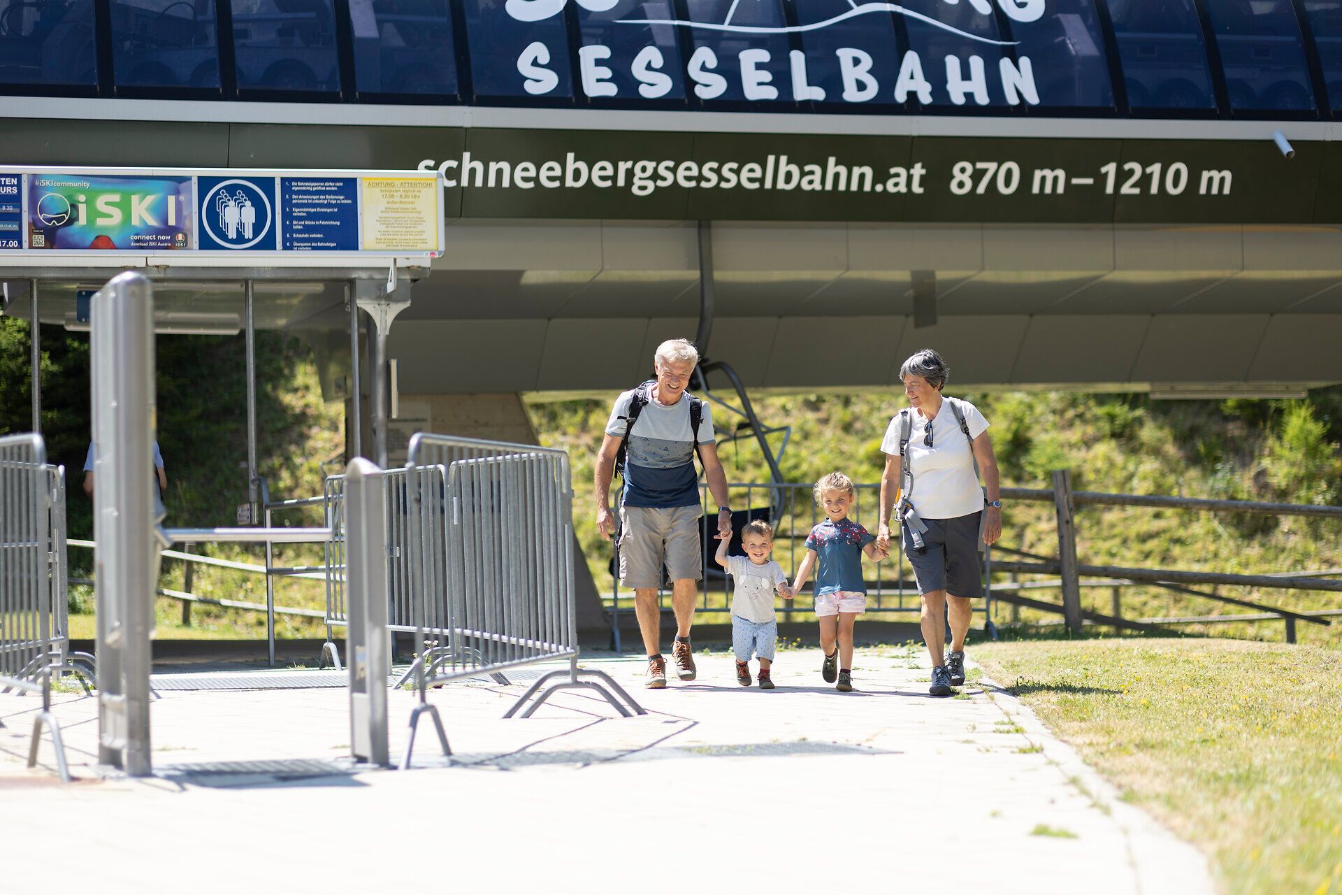 Eine Familie genießt einen sonnigen Tag in den Bergen, während sie fröhlich zur Sesselbahn schlendert. Die Kinder lachen und halten sich an den Händen, umgeben von der atemberaubenden Natur des Schneeberglands. Hier wird der Sommer in den Bergen zum unvergesslichen Erlebnis.