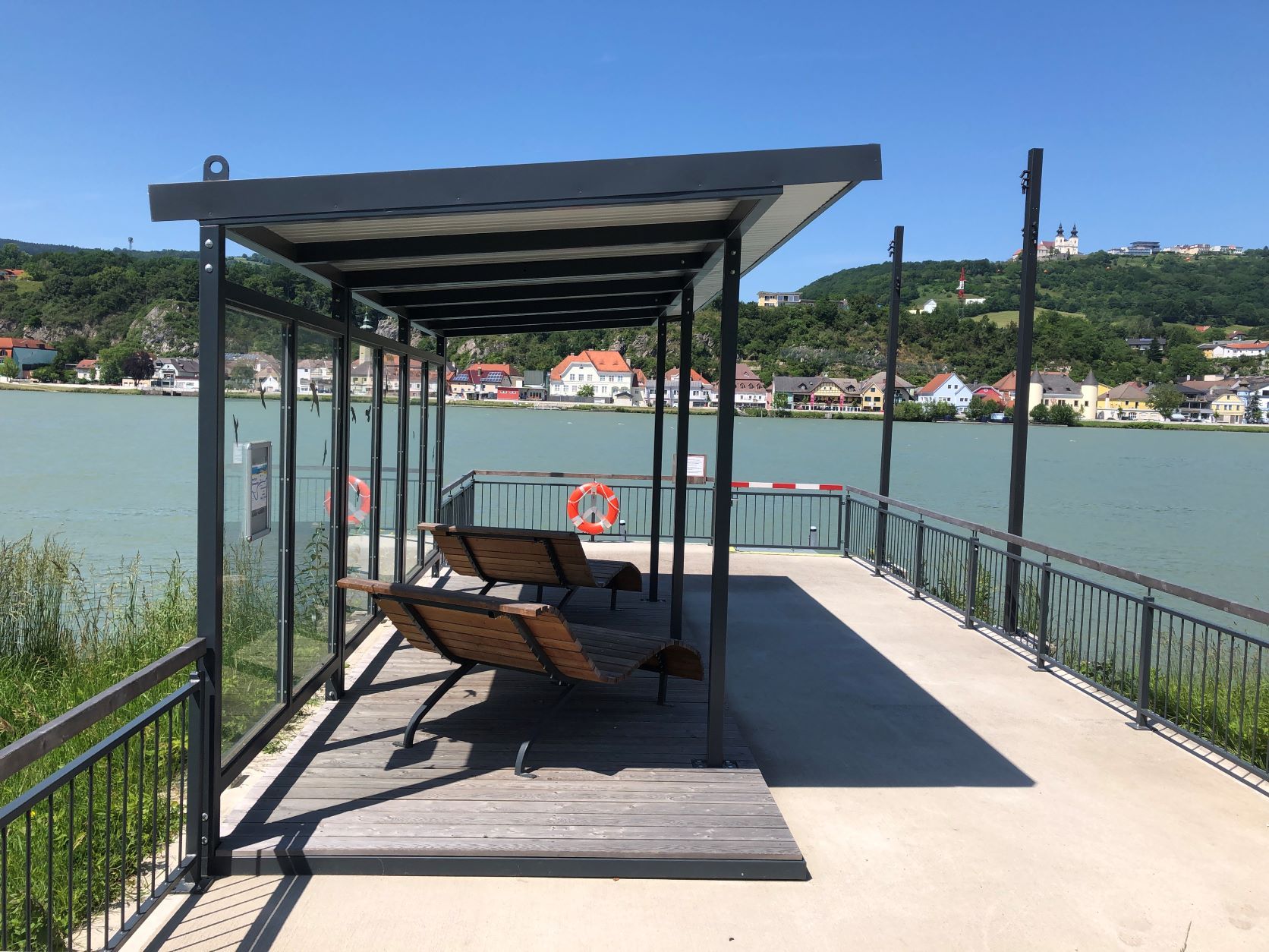 Anlegestelle mit Sitzbänken am Fluss, im Hintergrund eine Stadt und eine Basilika auf einem Hügel.
