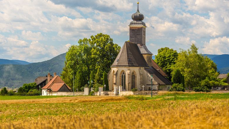 Kirche in Muthmannsdorf umgeben von Feldern und Bäumen, mit Bergen im Hintergrund.