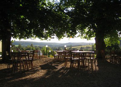 Ein schattiger Garten mit Holztischen und -stühlen unter großen Bäumen, mit Blick auf eine hügelige Landschaft im Hintergrund.