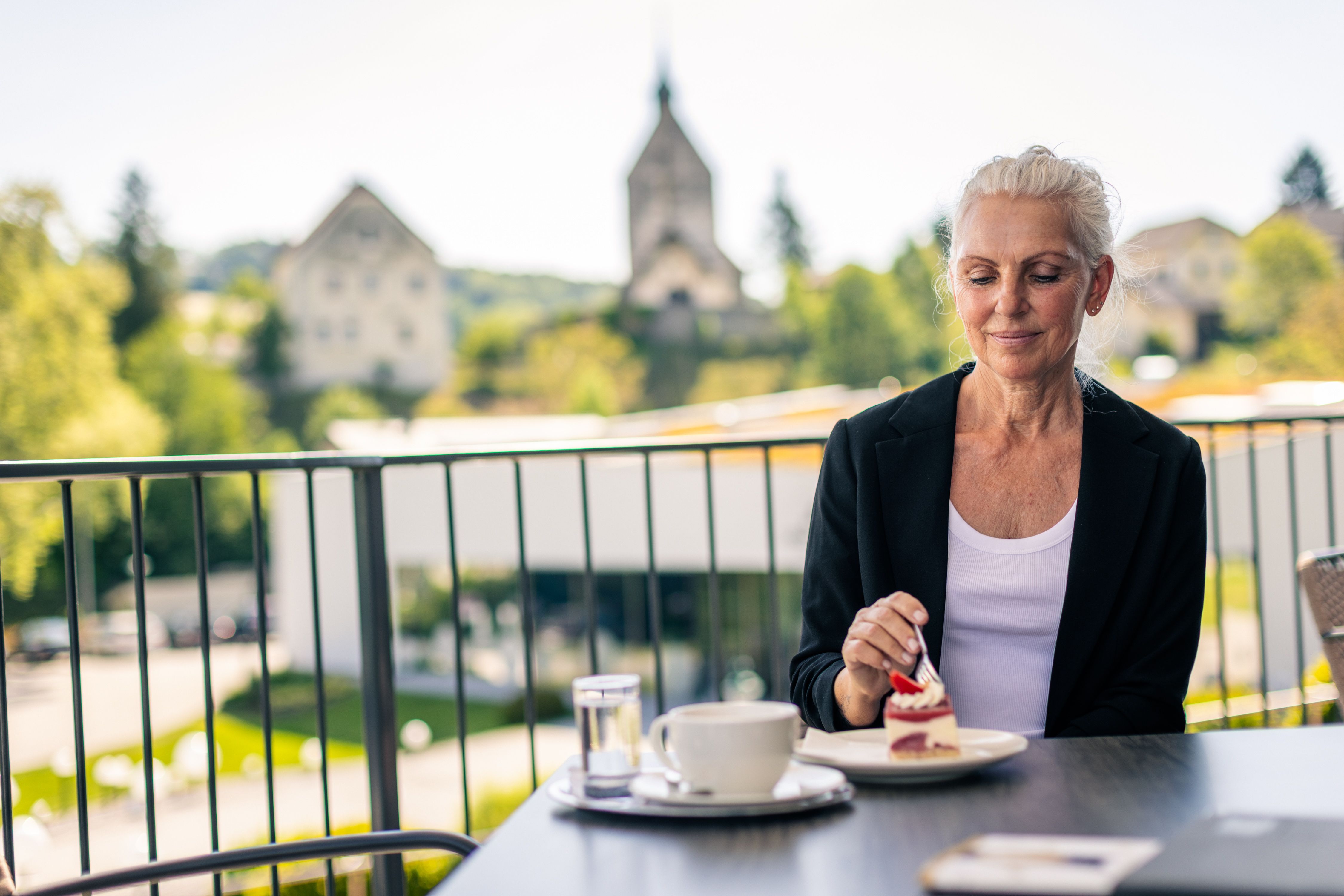 Eine Frau sitzt auf einer Terrasse und genießt ein Stück Kuchen mit einer Tasse Kaffee. Im Hintergrund ist die Wehrkirche Bad Schönau zu sehen.