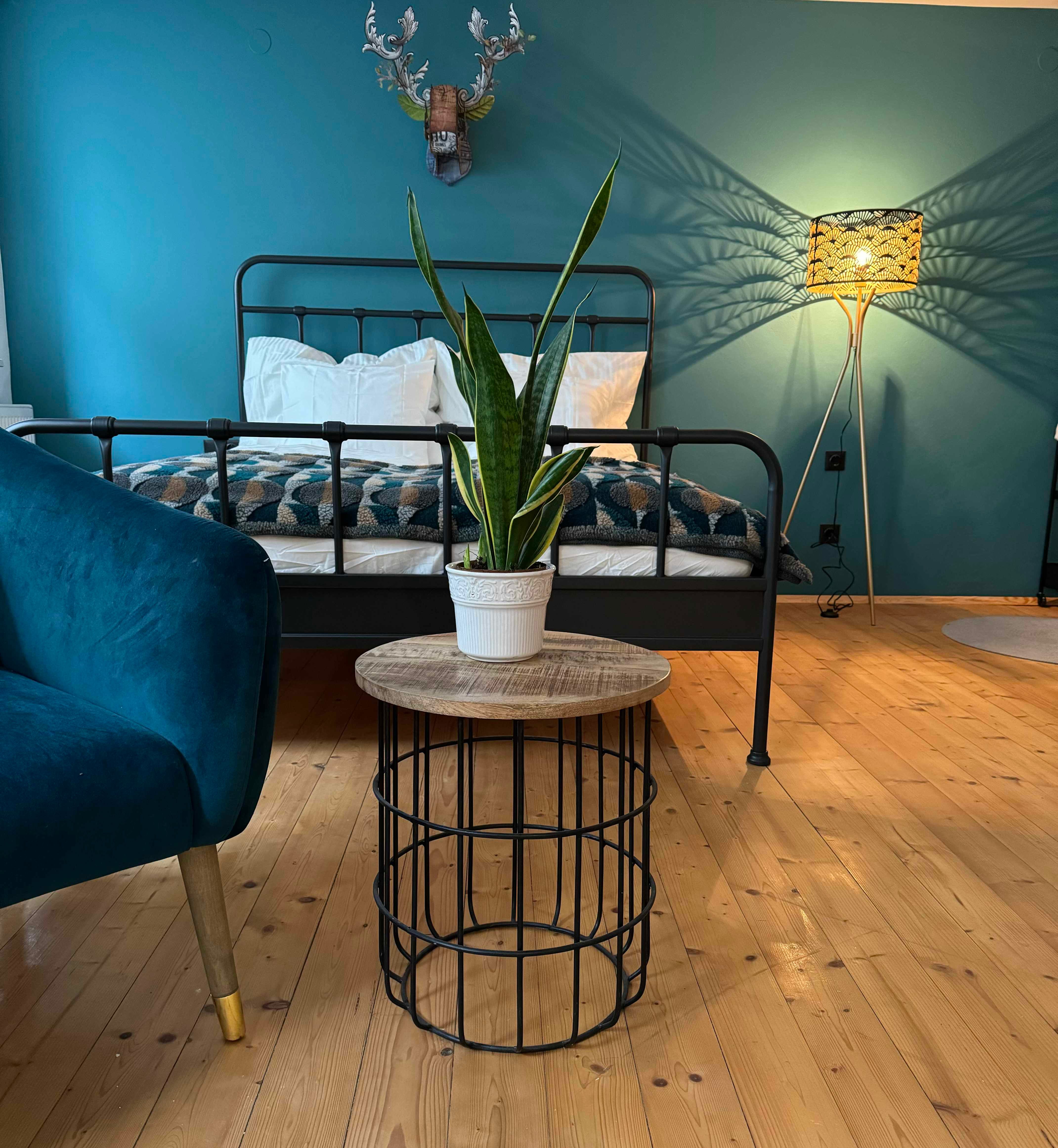 Schlafzimmer mit blauem Sessel, Metallbett, Pflanze auf Tisch und Stehlampe.