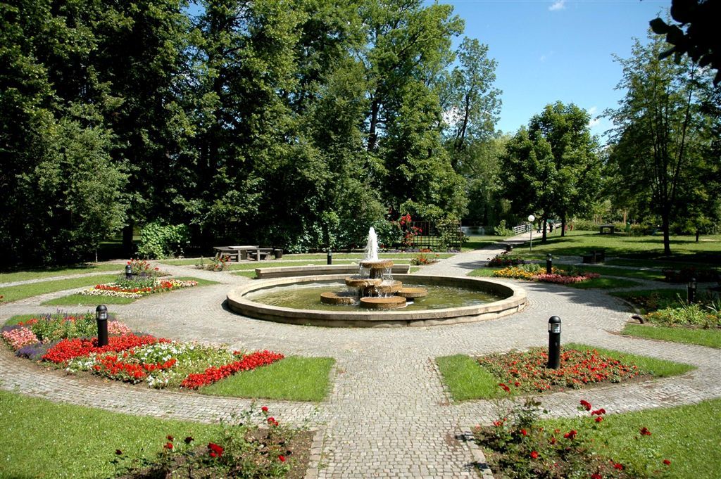 Ein gepflegter Park mit einem zentralen Brunnen, umgeben von Blumenbeeten und Bäumen.