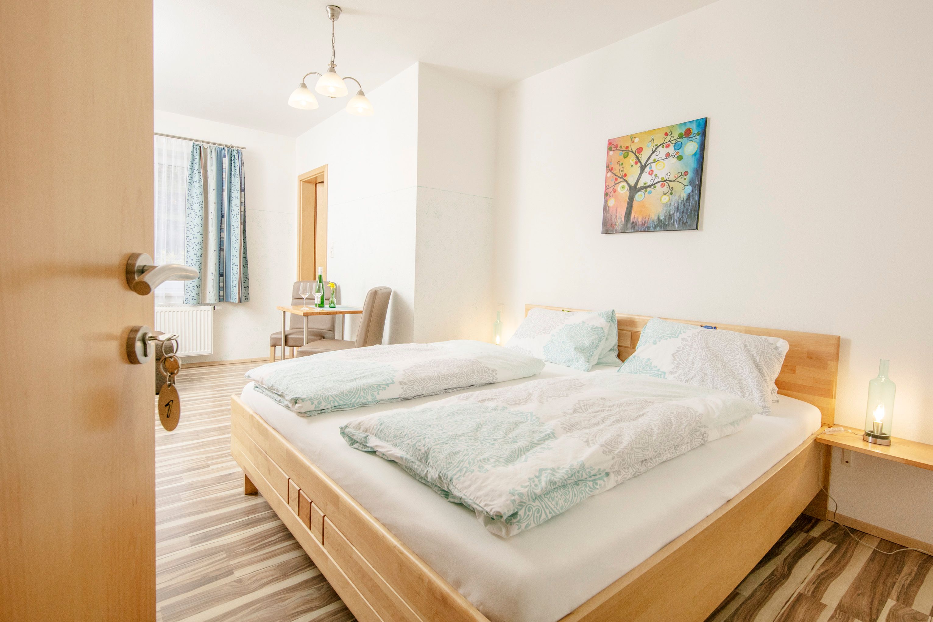 Ein helles Gästezimmer mit Doppelbett, Tisch, Stühlen und einem bunten Wandbild.