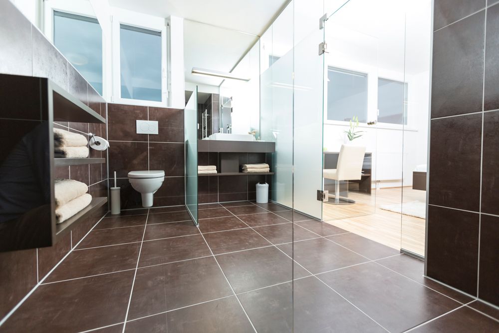 Modernes Badezimmer mit dunklen Fliesen, Glaswänden und weißen Sanitäreinrichtungen.