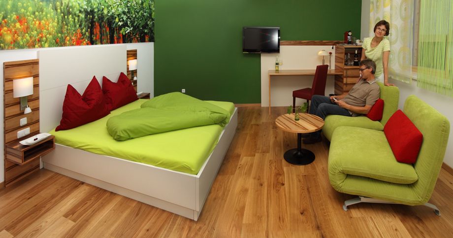 Ein modernes Hotelzimmer mit grünem und rotem Dekor, einem Doppelbett, einem Sofa und einem kleinen Tisch. Zwei Personen sind im Raum zu sehen.