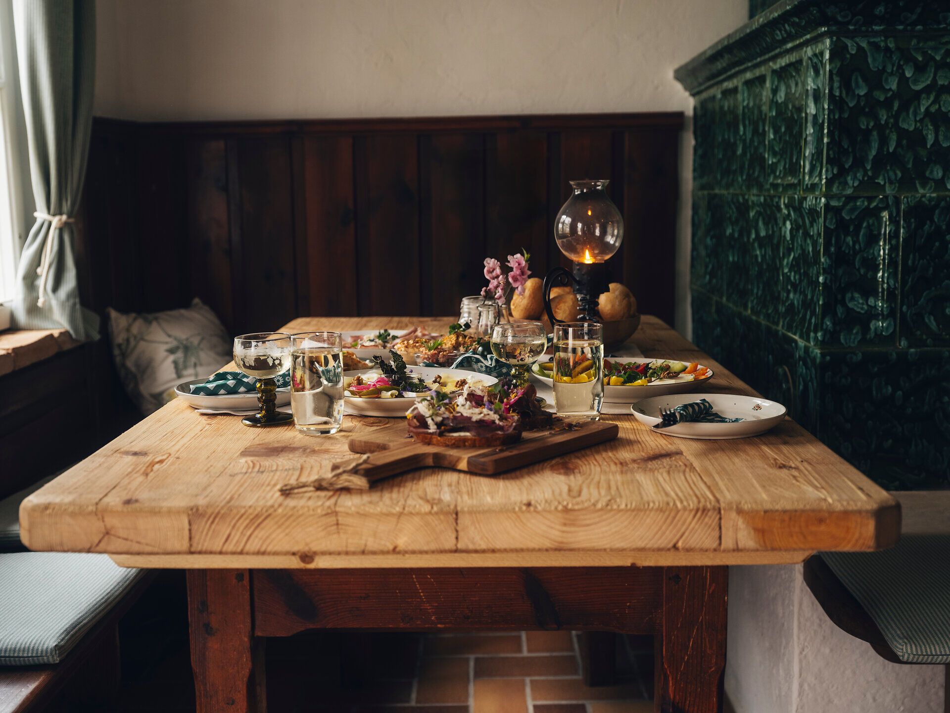 Rustikaler Holztisch mit angerichteten Speisen, Brot, Gläsern und Blumen in einer gemütlichen Stube; daneben ein grüner Kachelofen und Holzvertäfelung.