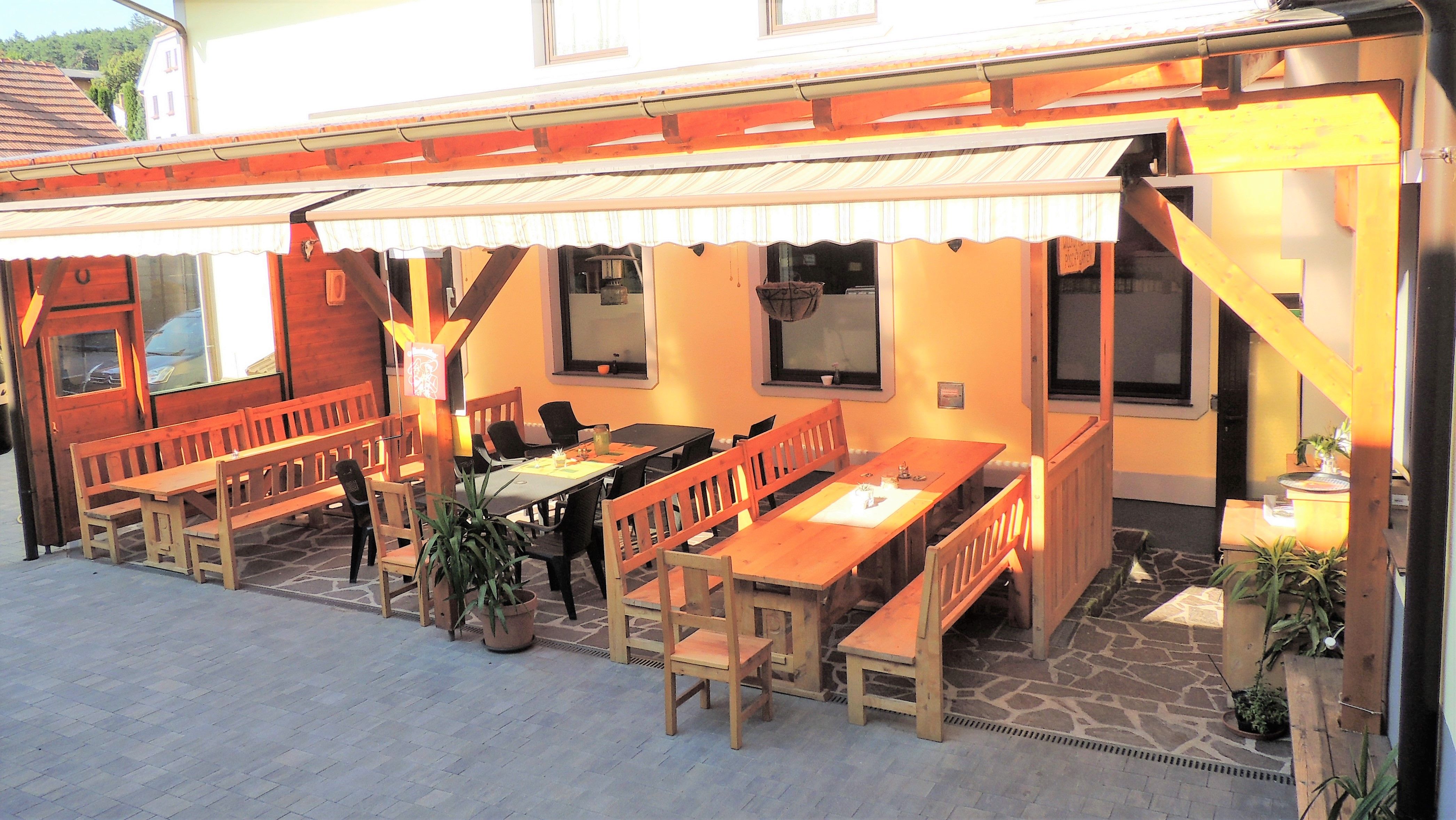 Ein gemütlicher Gastgarten mit Holzbänken und Tischen unter einer Markise.