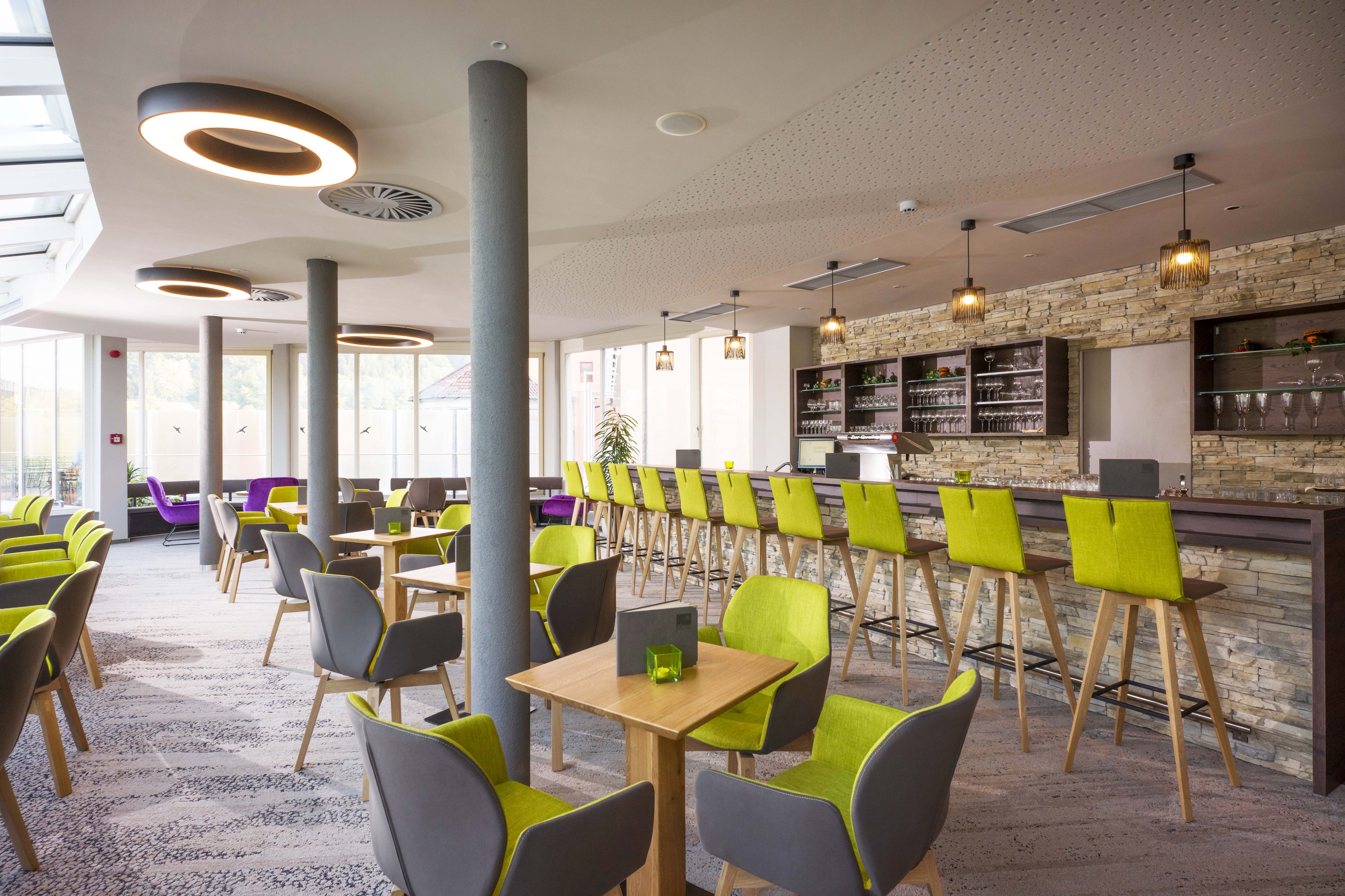 Modernes Café mit grünen und grauen Stühlen, Holztischen und einer langen Bar mit Steinwand.