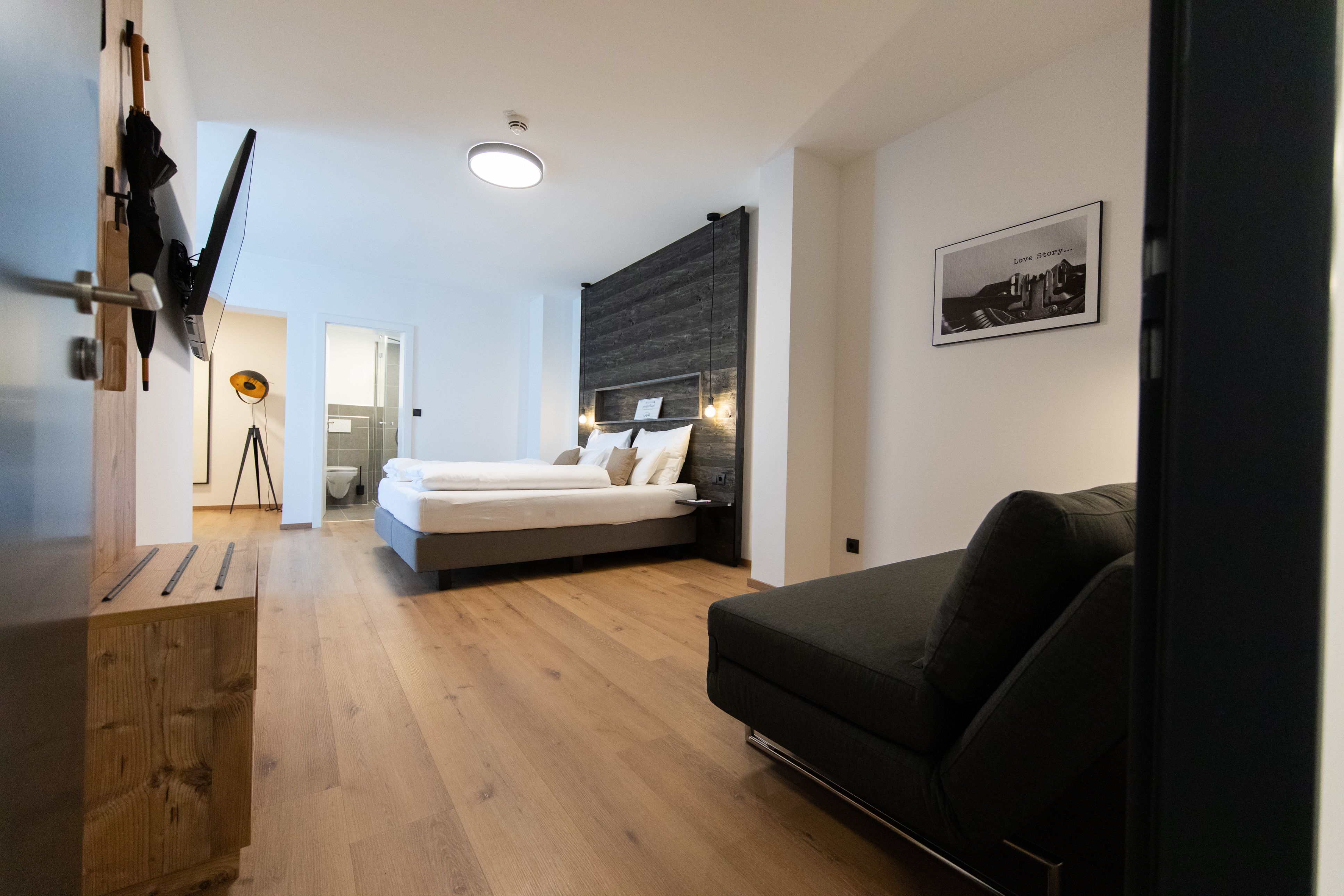 Modern eingerichtetes Hotelzimmer mit Doppelbett, Sofa und Flachbildfernseher. Helle Holzböden und ein Badezimmer im Hintergrund.