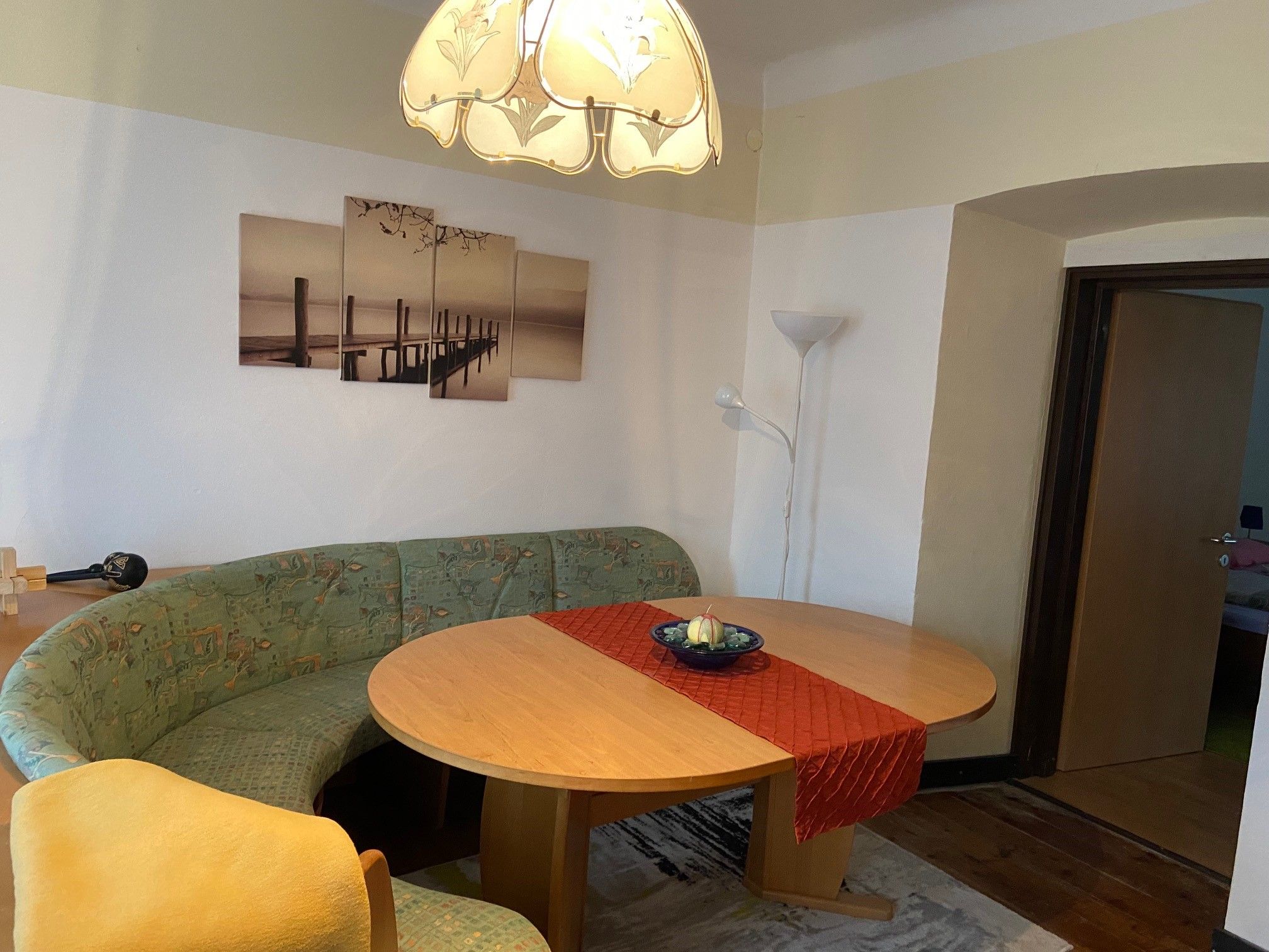 Ein Wohnzimmer mit einem ovalen Holztisch, einer grünen Eckbank, einem roten Tischläufer und einem Bild an der Wand. Eine Stehlampe steht in der Ecke.