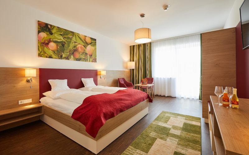 Modern eingerichtetes Hotelzimmer mit Doppelbett, roter Decke, Holzmöbeln und großem Fenster. An der Wand hängt ein Bild mit Früchten.