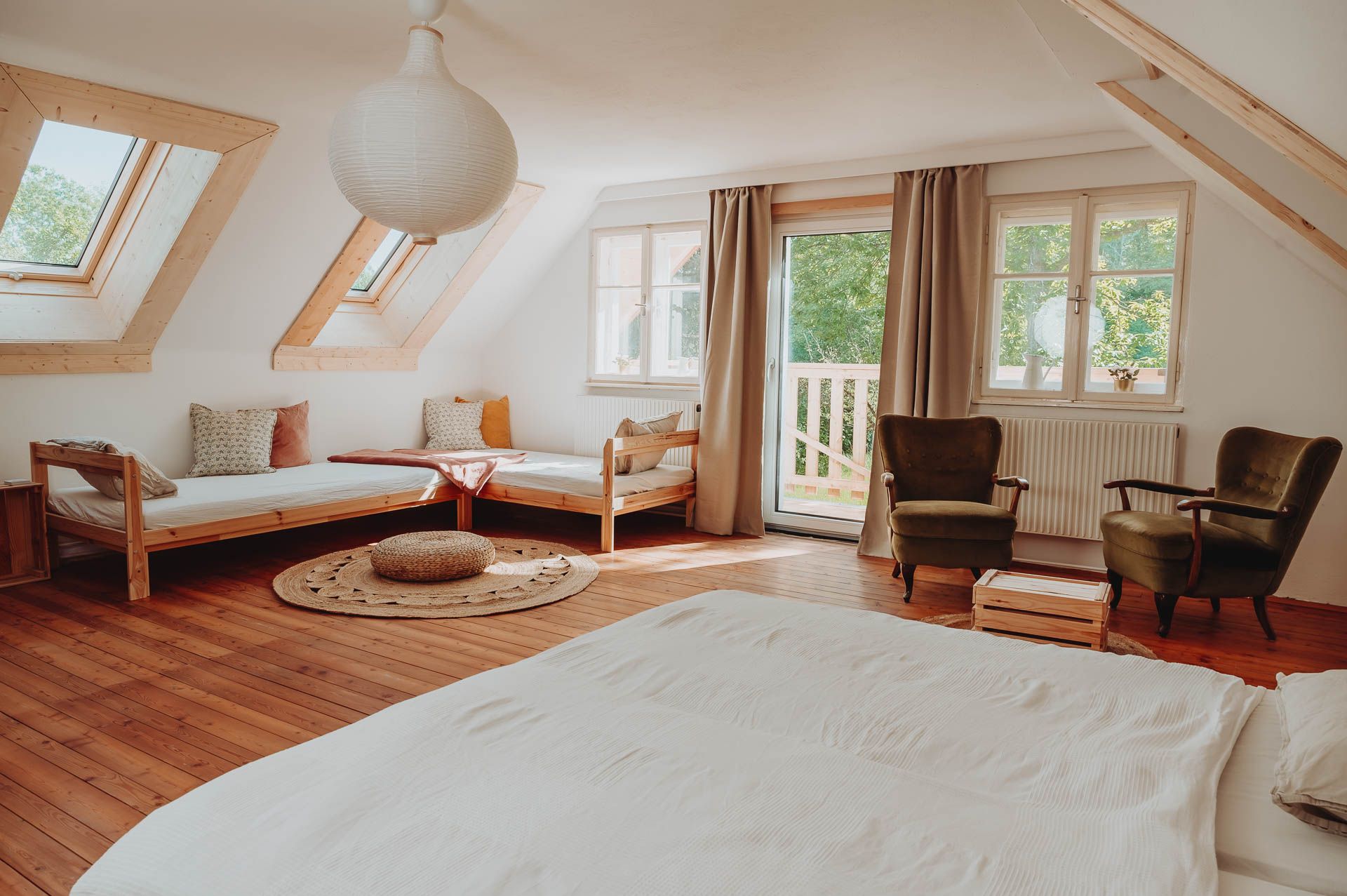 Helles Mehrbettzimmer mit Holzboden, zwei Betten, zwei Sesseln und großen Fenstern.