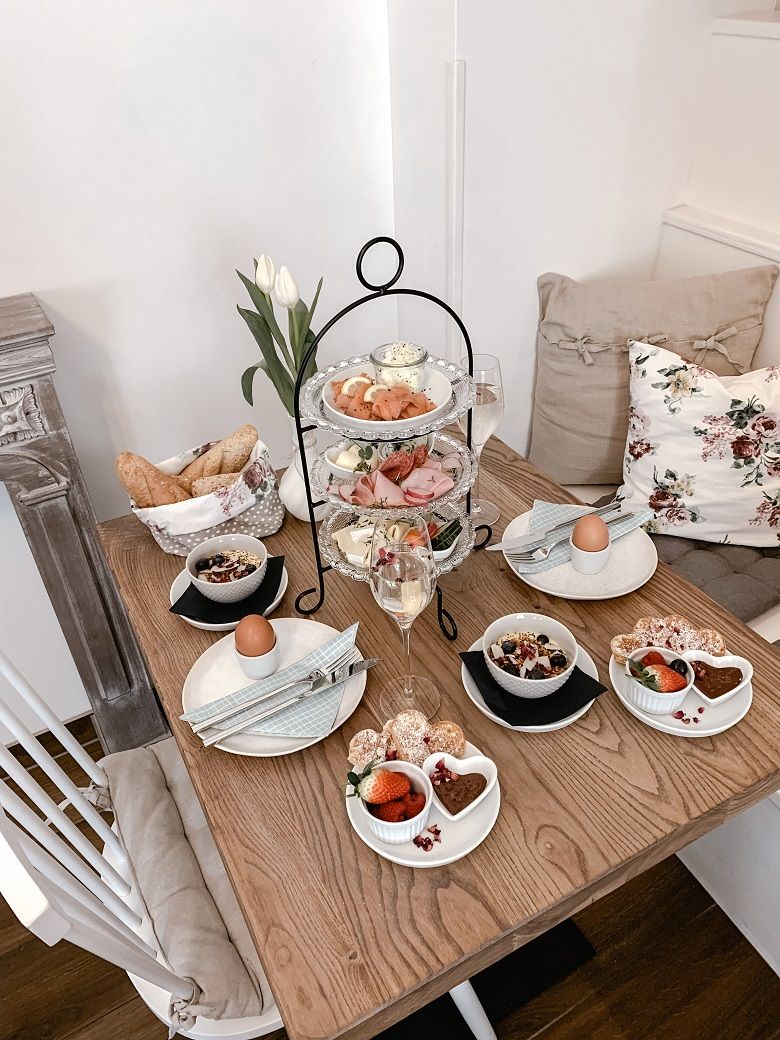 Ein gedeckter Frühstückstisch mit Etagere, Brötchen, Eiern, Müsli und Obst. Dekoriert mit Tulpen und Kissen.
