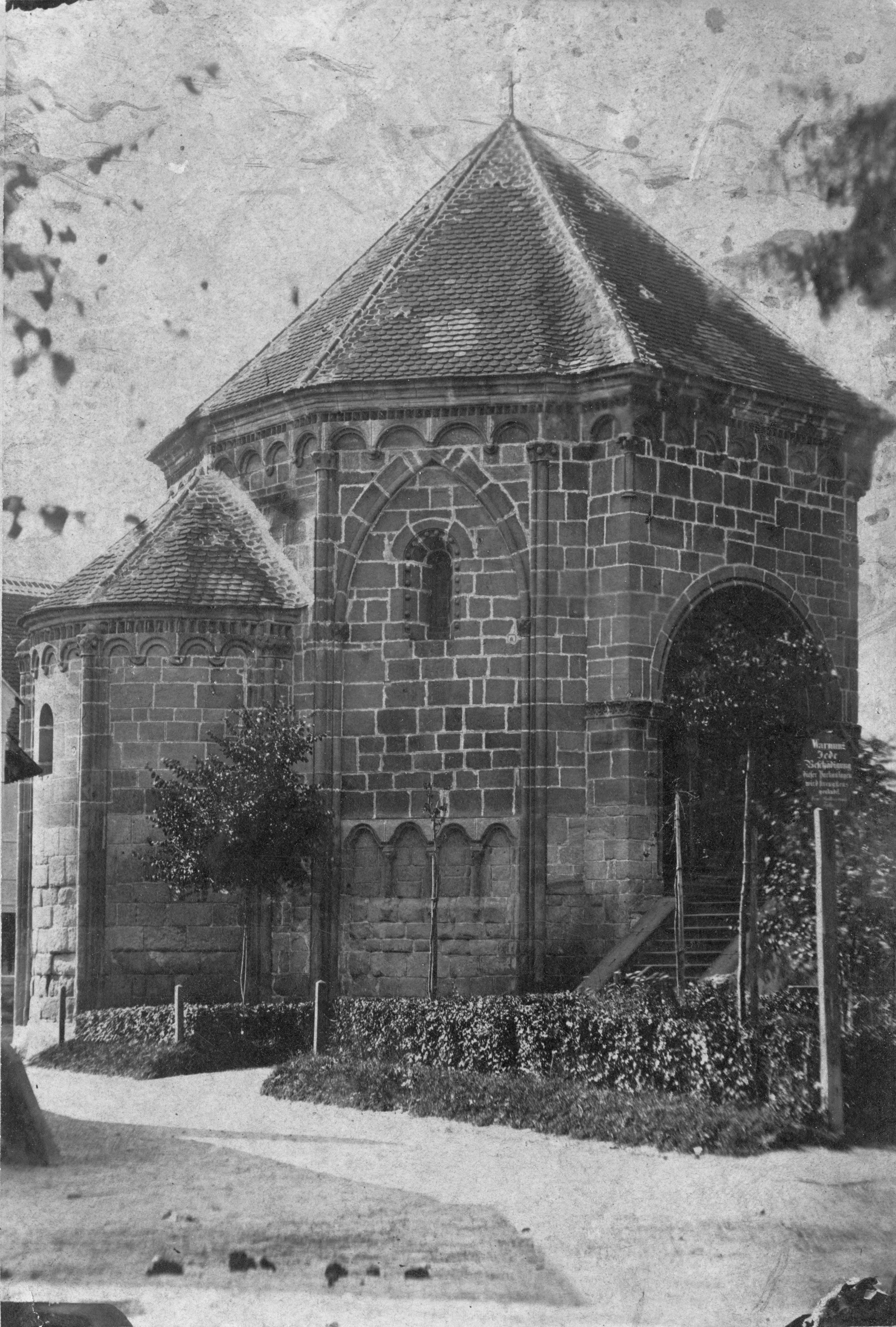 Historisches Foto eines spätromanischen Karners mit Rundbogenfenstern und Ziegeldach, umgeben von Bäumen und einem Weg.