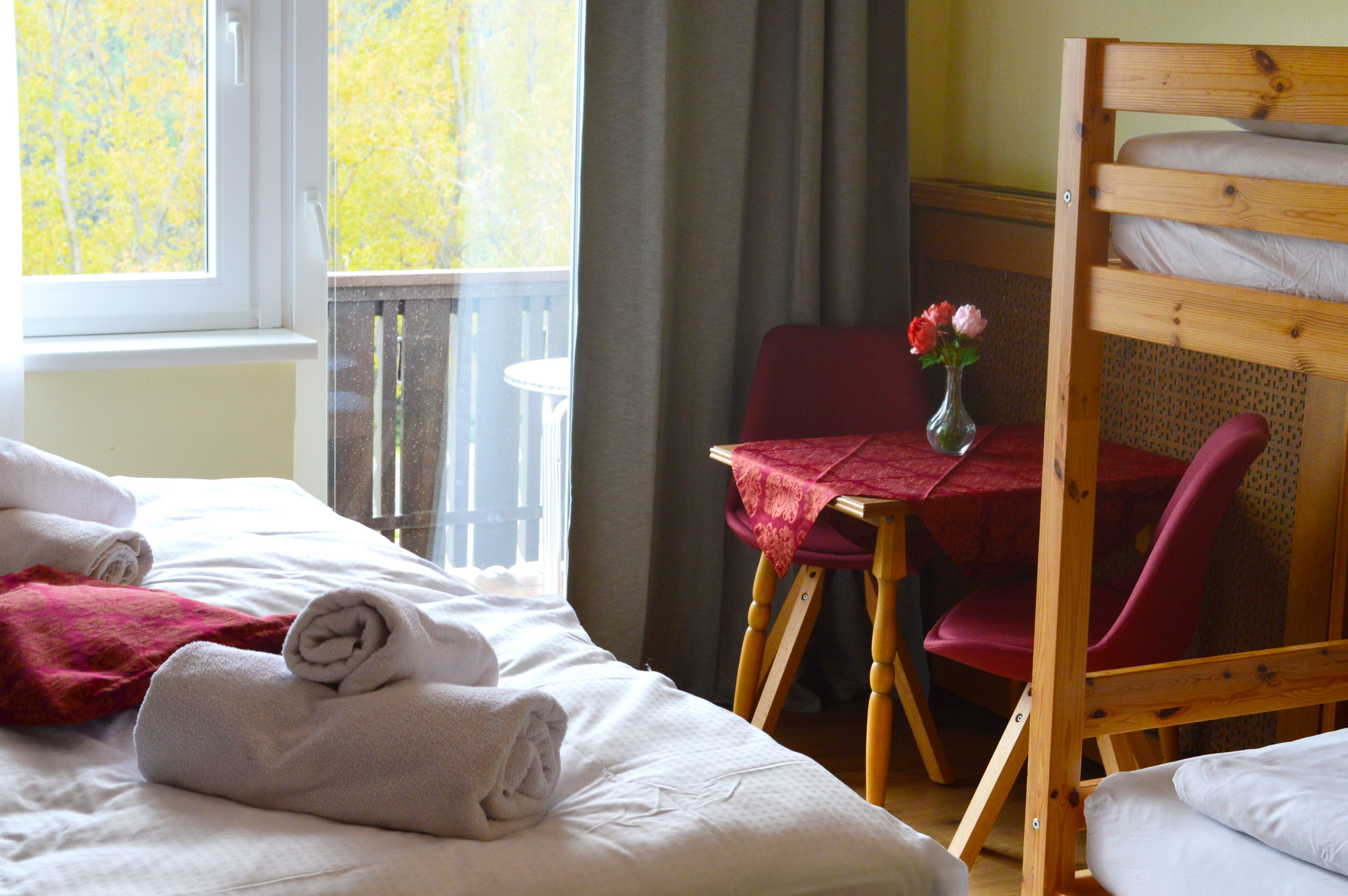 Ein gemütliches Zimmer mit einem Bett, einem Tisch mit roten Stühlen und einem Blumenstrauß. Ein Fenster zeigt einen Balkon und herbstliche Bäume.