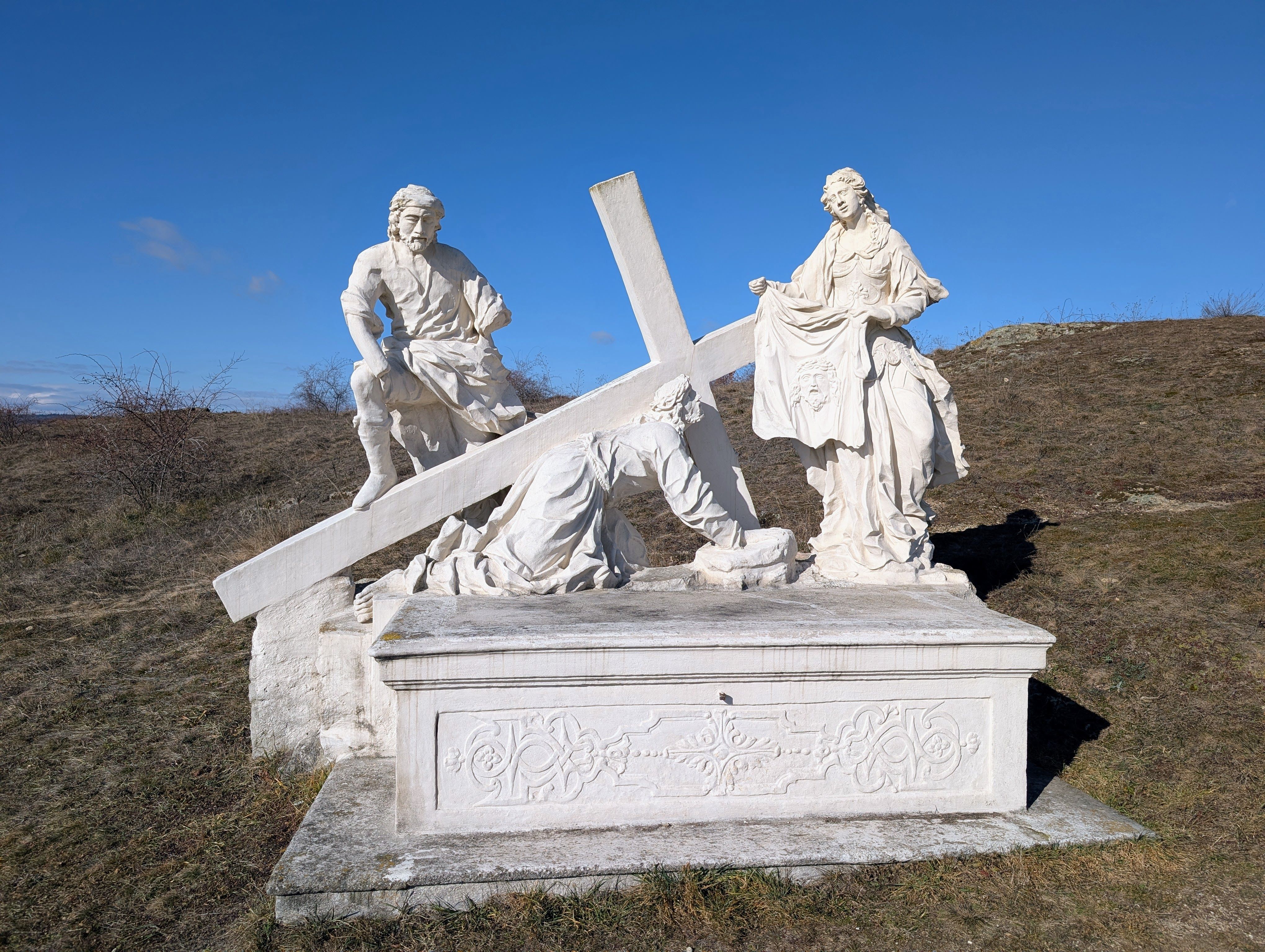 Steinskulptur einer religiösen Szene mit drei Figuren und einem Kreuz auf einem Hügel.
