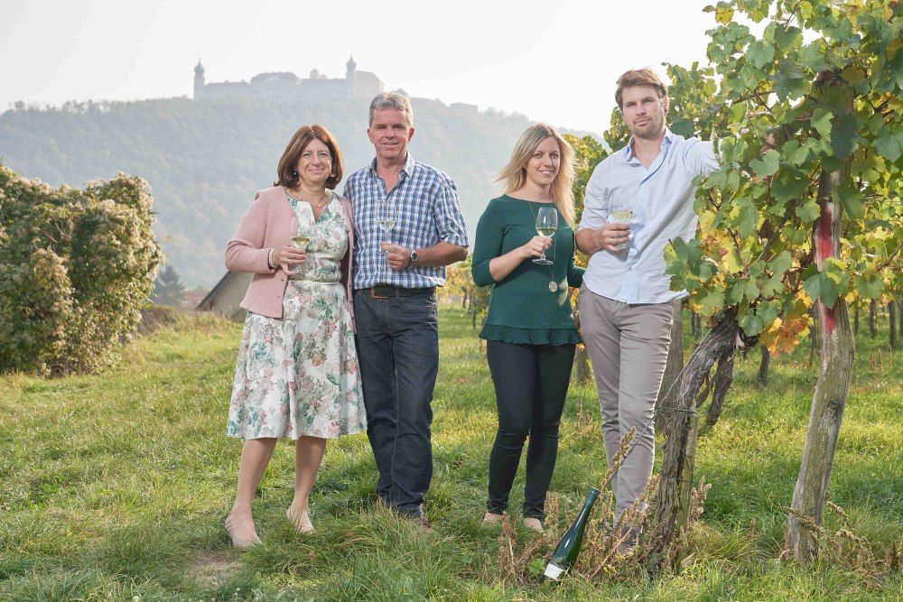 Vier Personen stehen in einem Weinberg mit Weingläsern in der Hand, im Hintergrund ist eine Burg auf einem Hügel zu sehen.