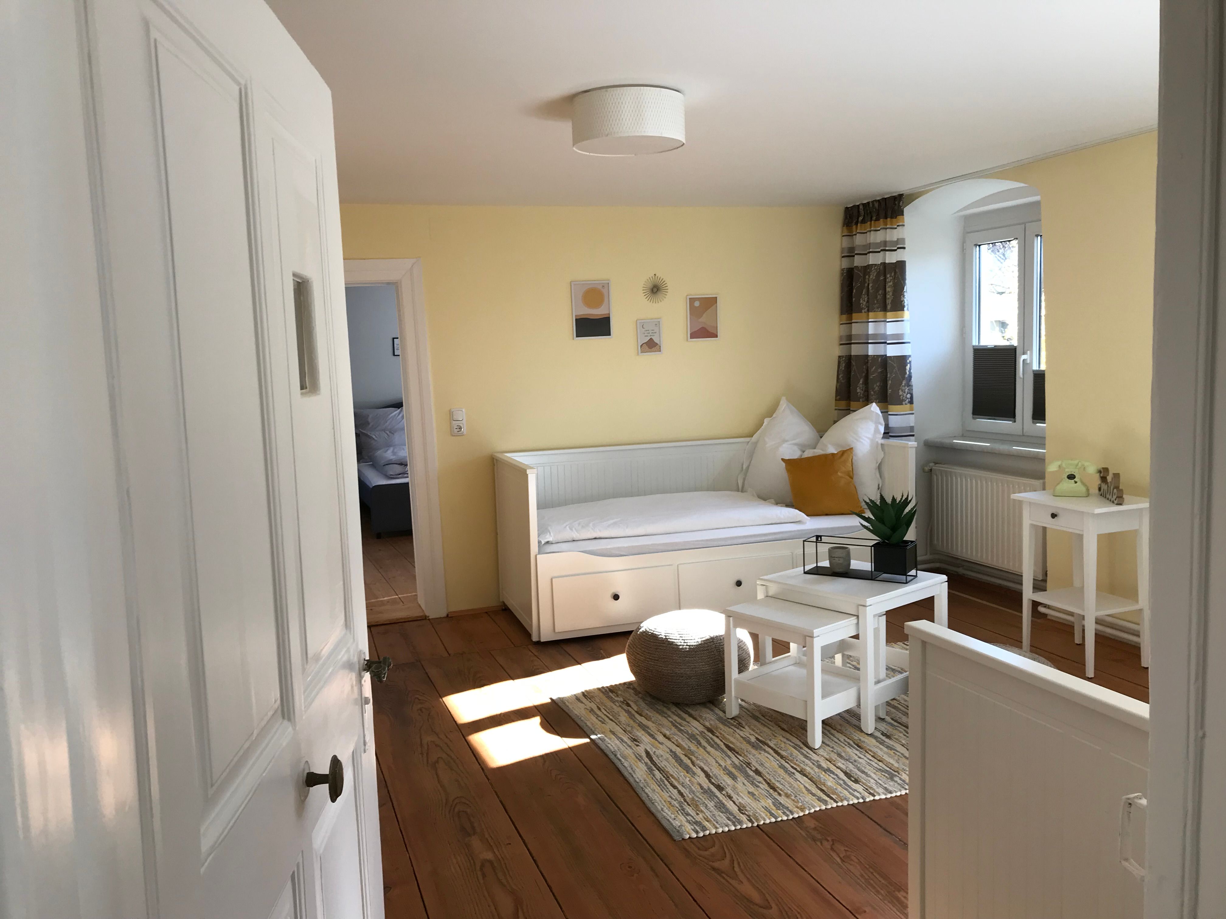 Heller Raum mit gelben Wänden, einem weißen Tagesbett, kleinem Tisch und Pflanzen.