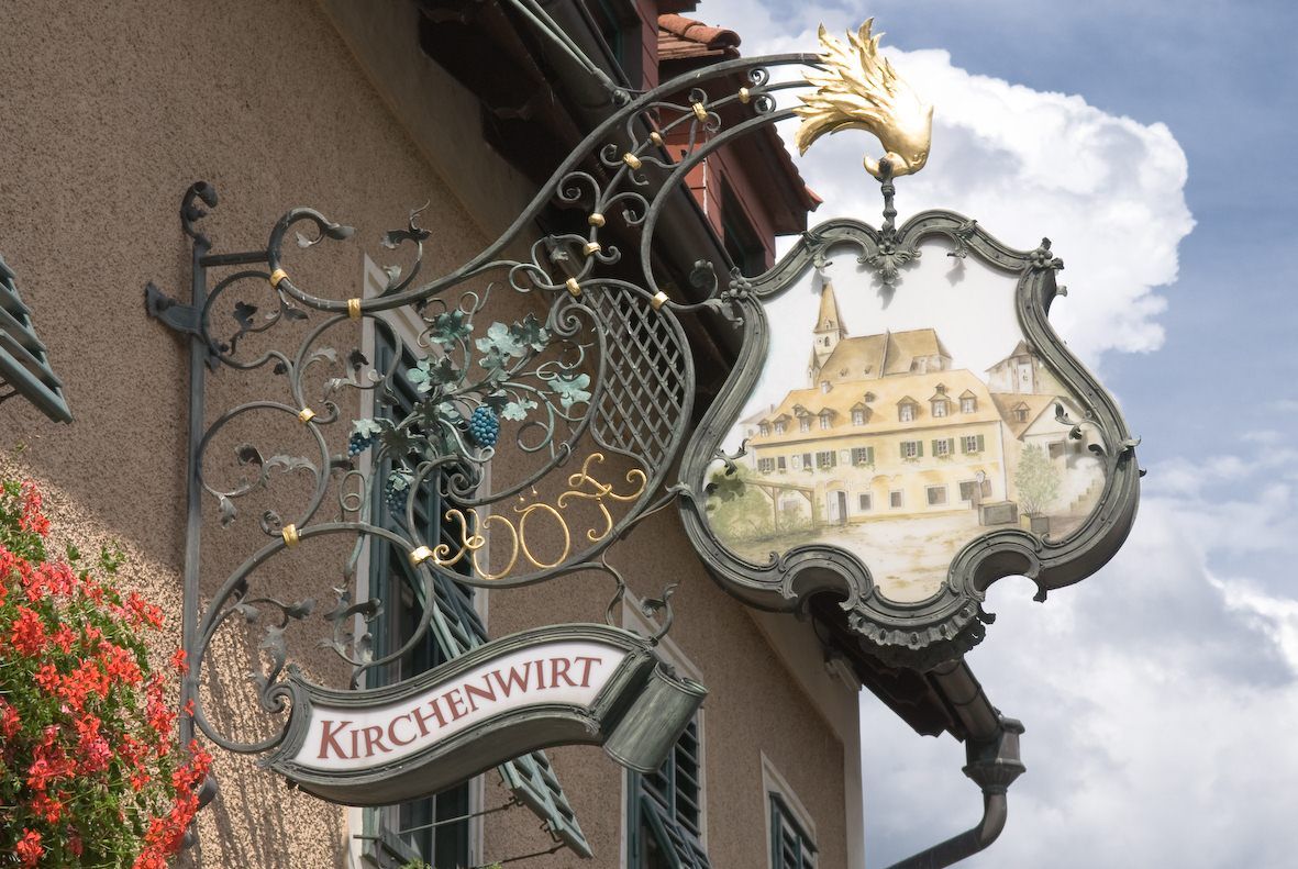 Ein kunstvoll gestaltetes Wirtshausschild mit der Aufschrift 'Kirchenwirt' und einem Bild eines Gebäudes mit Kirchturm.