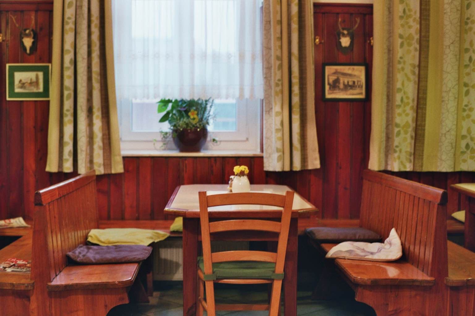 Gemütliche Gaststube mit Holzmöbeln, Tisch, Stühlen und Bank, Fenster mit Vorhängen, Pflanzen und Wanddekorationen.